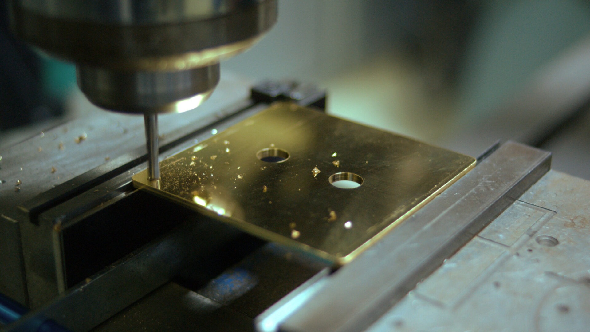 A drill precisely machines a metallic, gold-colored plate on a workshop table, creating two holes. Small metal shavings are scattered around the area.