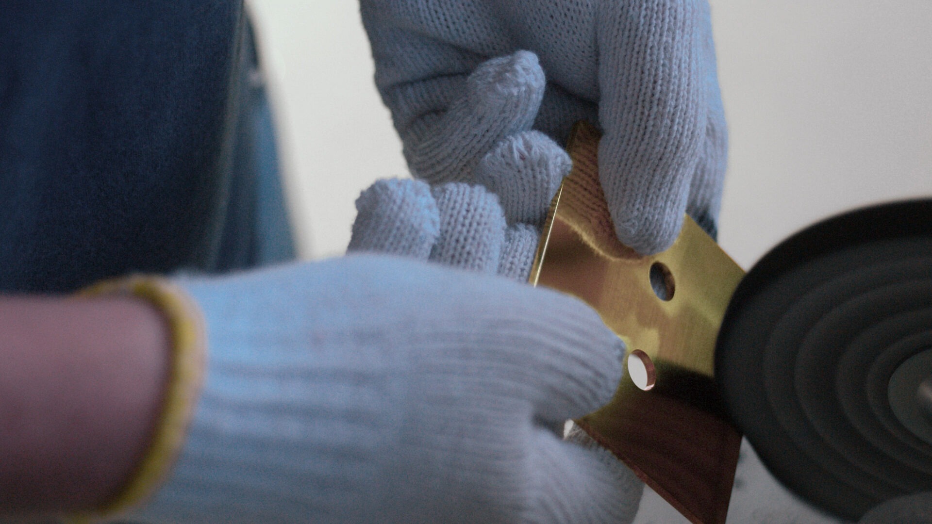 A person in gloves holds a shiny metallic object near a power tool, indicating a detailed crafting or polishing process.