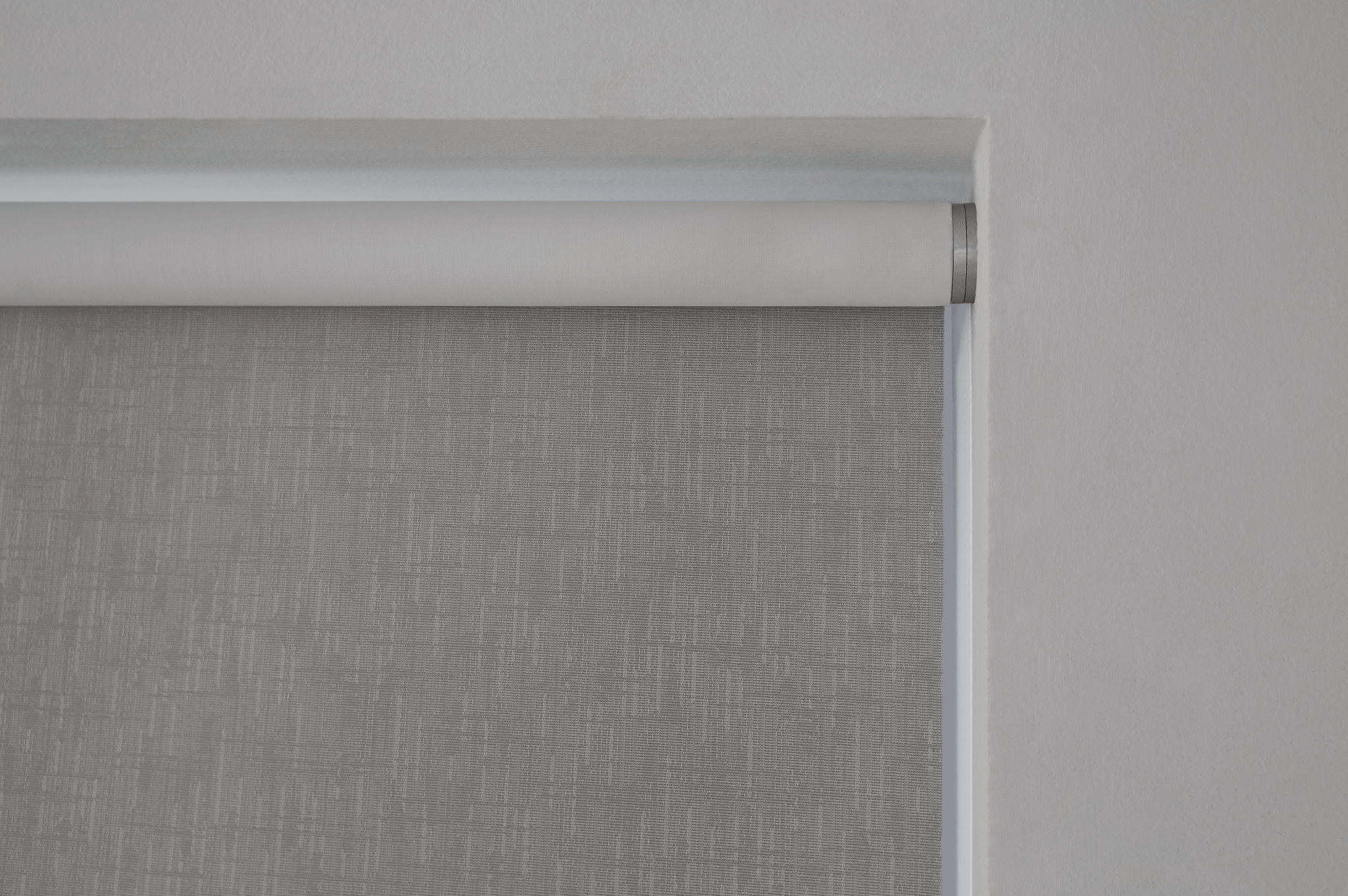 A gray roller blind partially unfurled against a white wall, showcasing a minimalist, textured design without any visible landmarks or historical buildings.