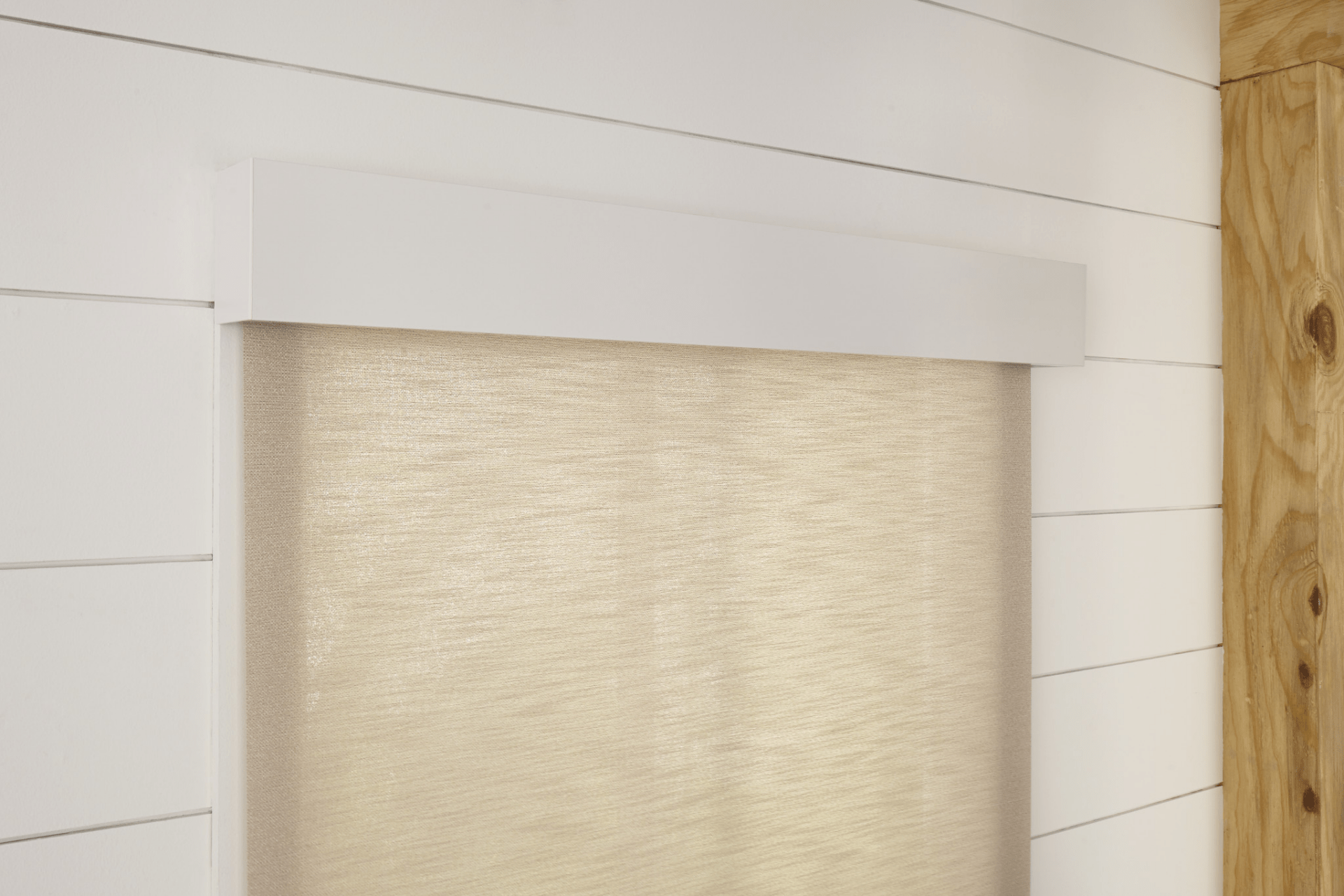 A window with a beige roller shade against a white, wooden shiplap wall. The shades are neatly rolled and the wall is simple.