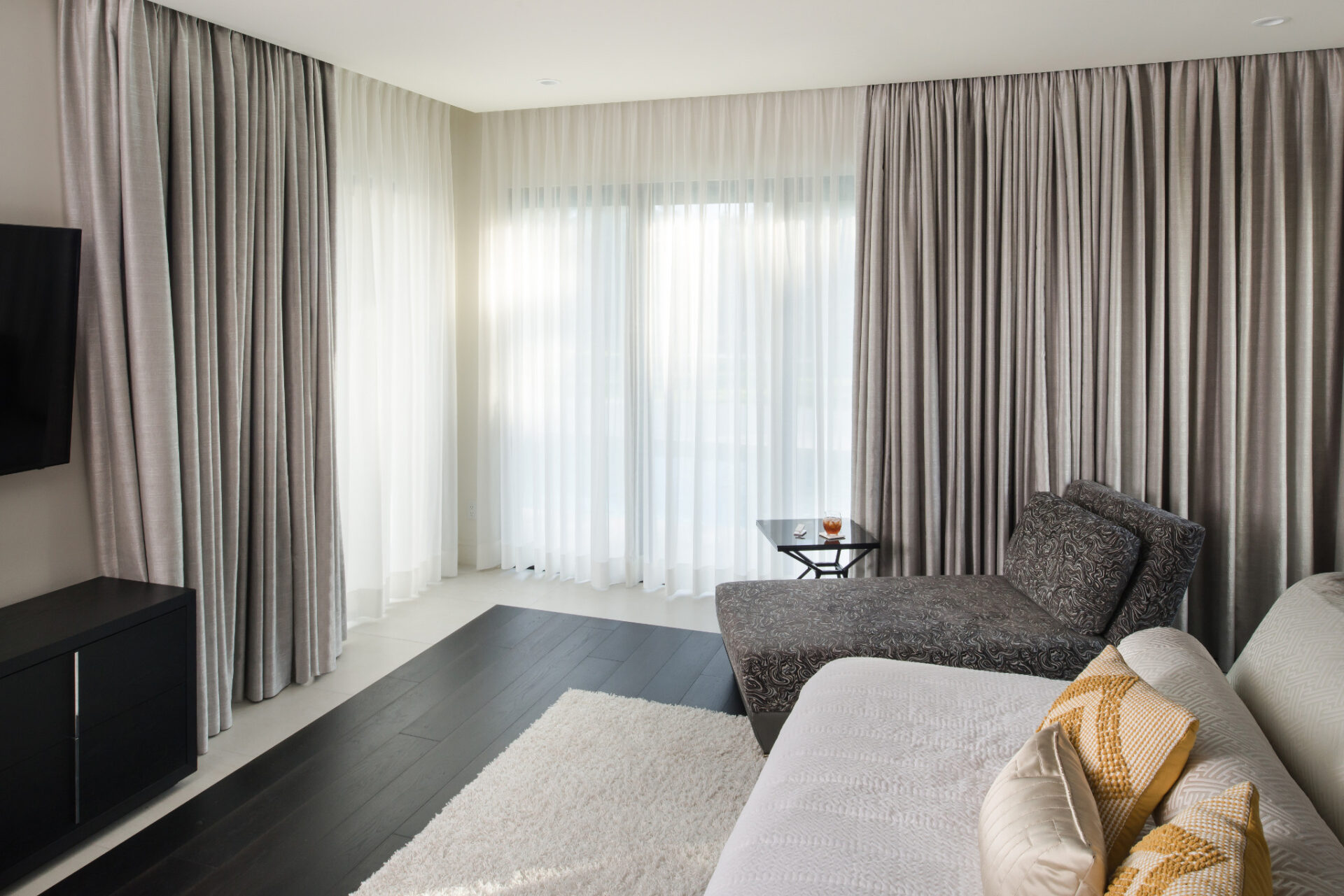 A modern living room features elegant, gray curtains, a cozy chaise lounge, and a white rug on dark hardwood floors, creating a serene atmosphere.