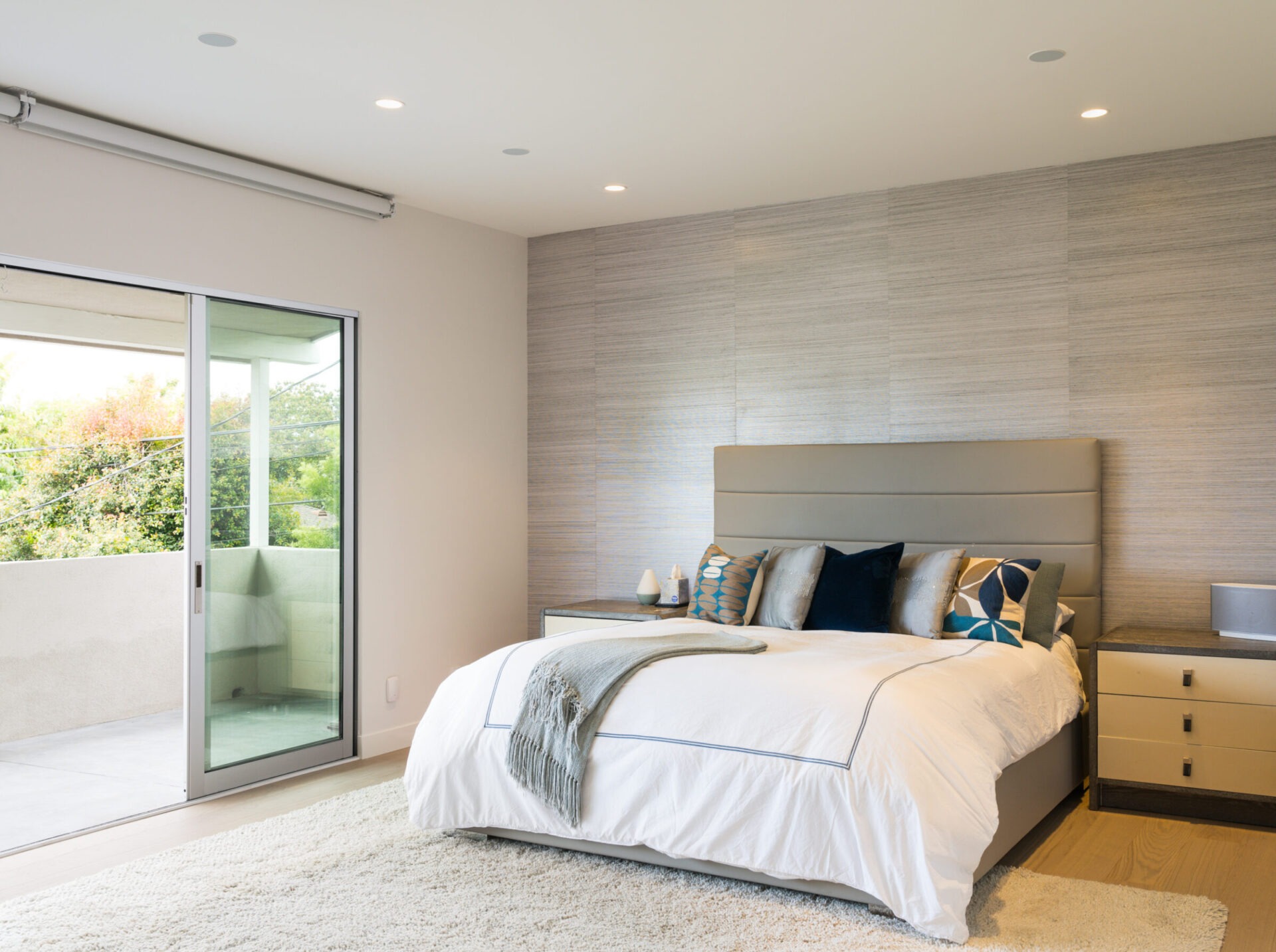 Modern bedroom with large bed, plush rug, and sliding glass doors leading to a balcony. Neutral tones and soft lighting create a cozy atmosphere.