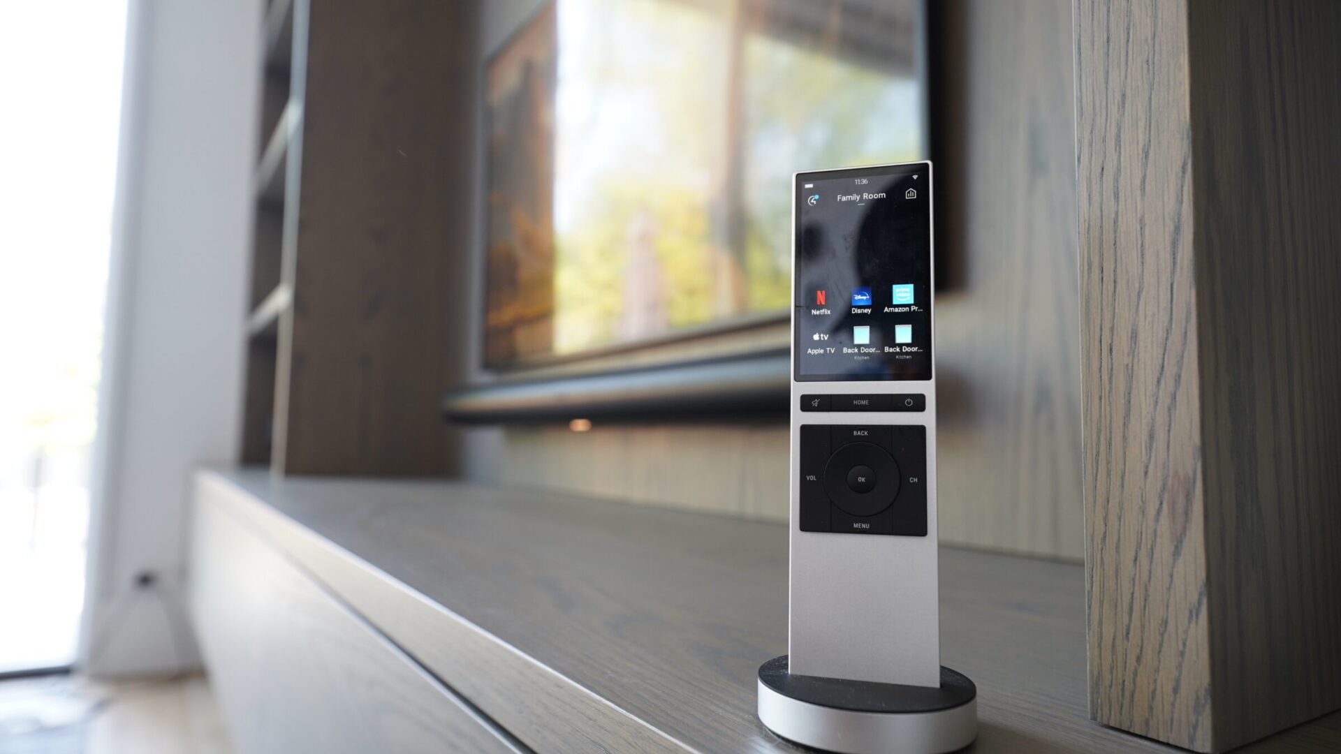 A sleek remote control device on a wooden shelf, featuring a modern design and digital display, with a blurred background of a living room.