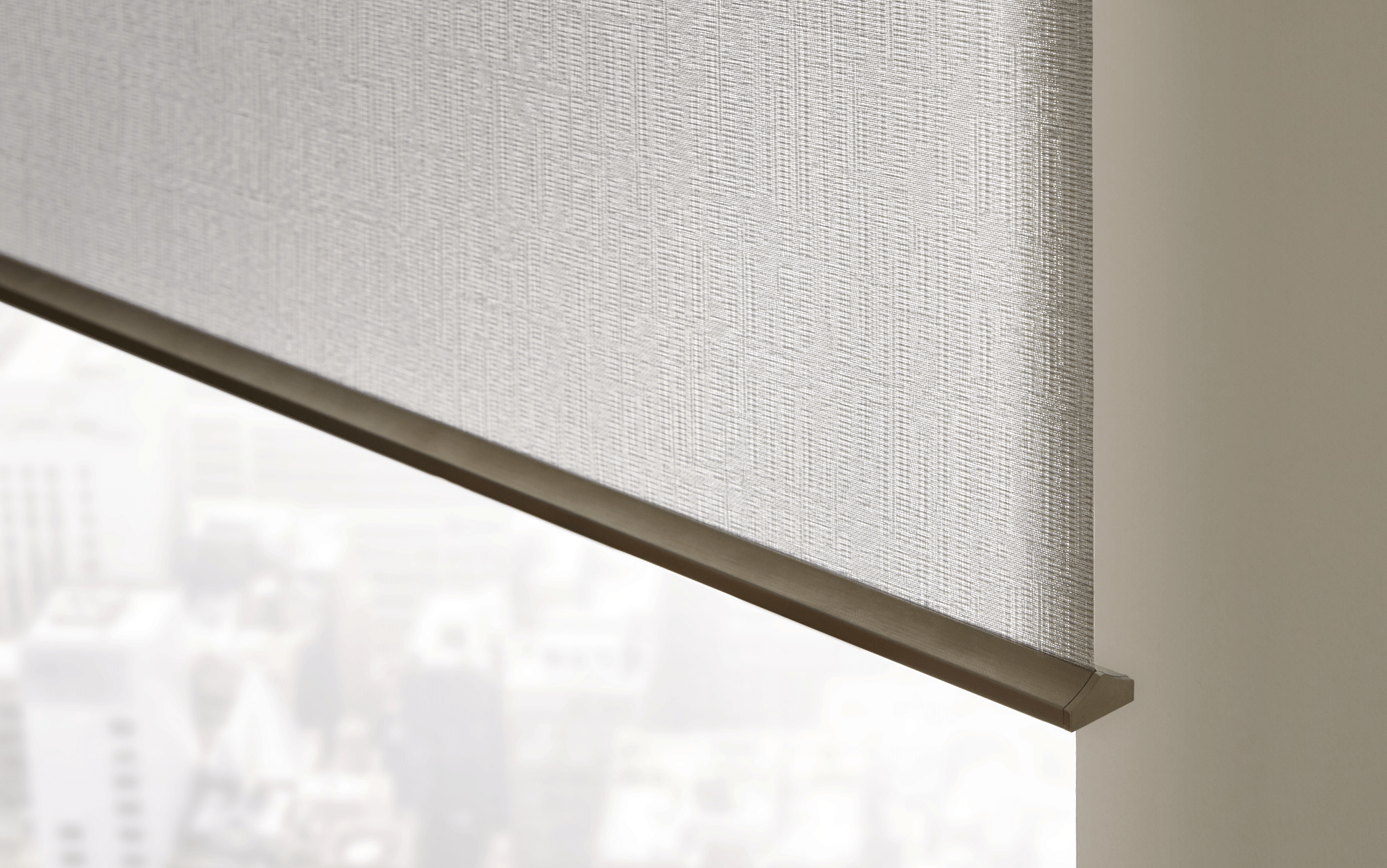 A close-up of a fabric roller blind in a high-rise building, partially concealing a blurred cityscape with tall buildings in the background.