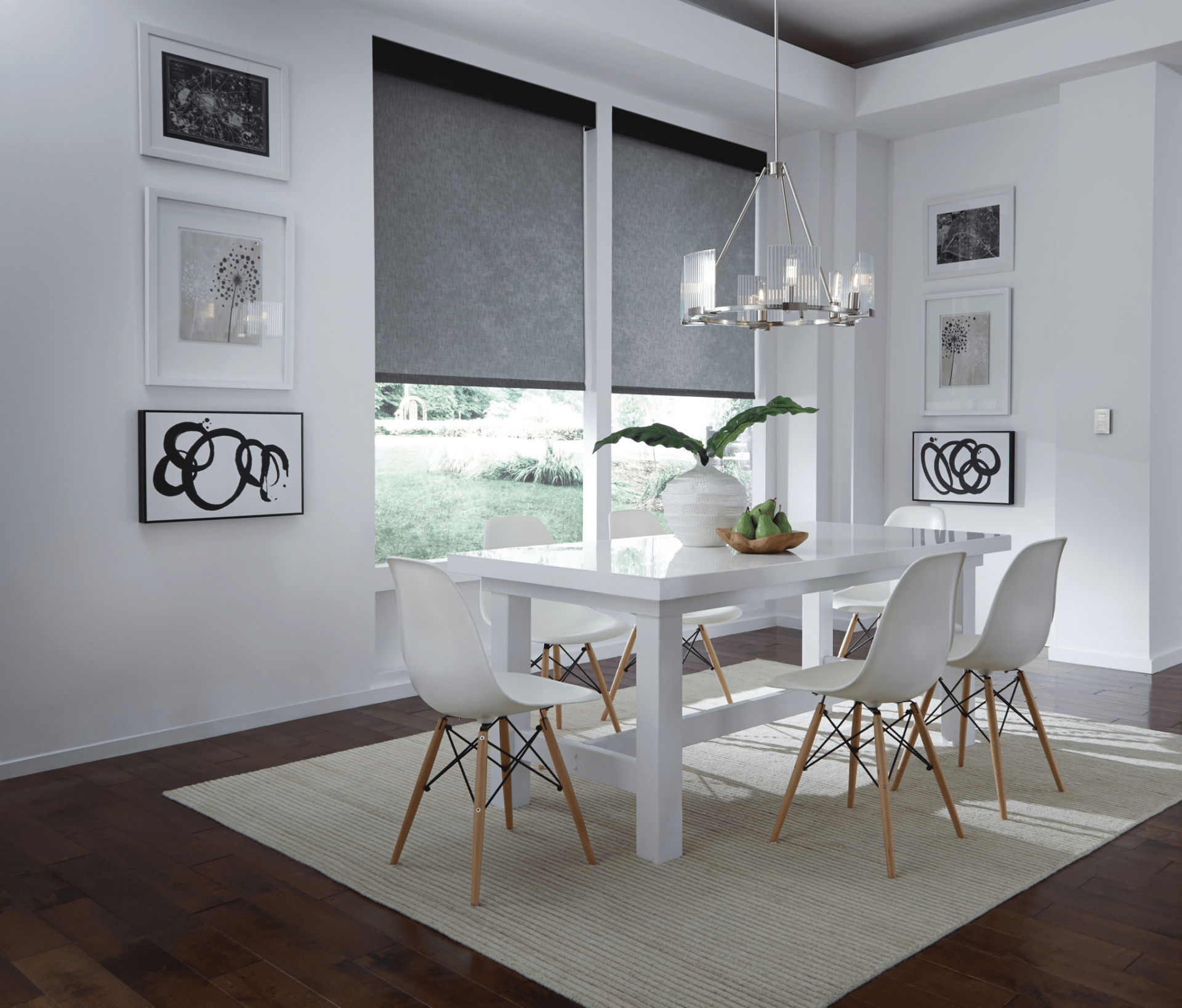 Modern dining room with white table, four chairs, abstract art, and chandelier. Large windows with gray blinds. Hardwood floor and textured rug.