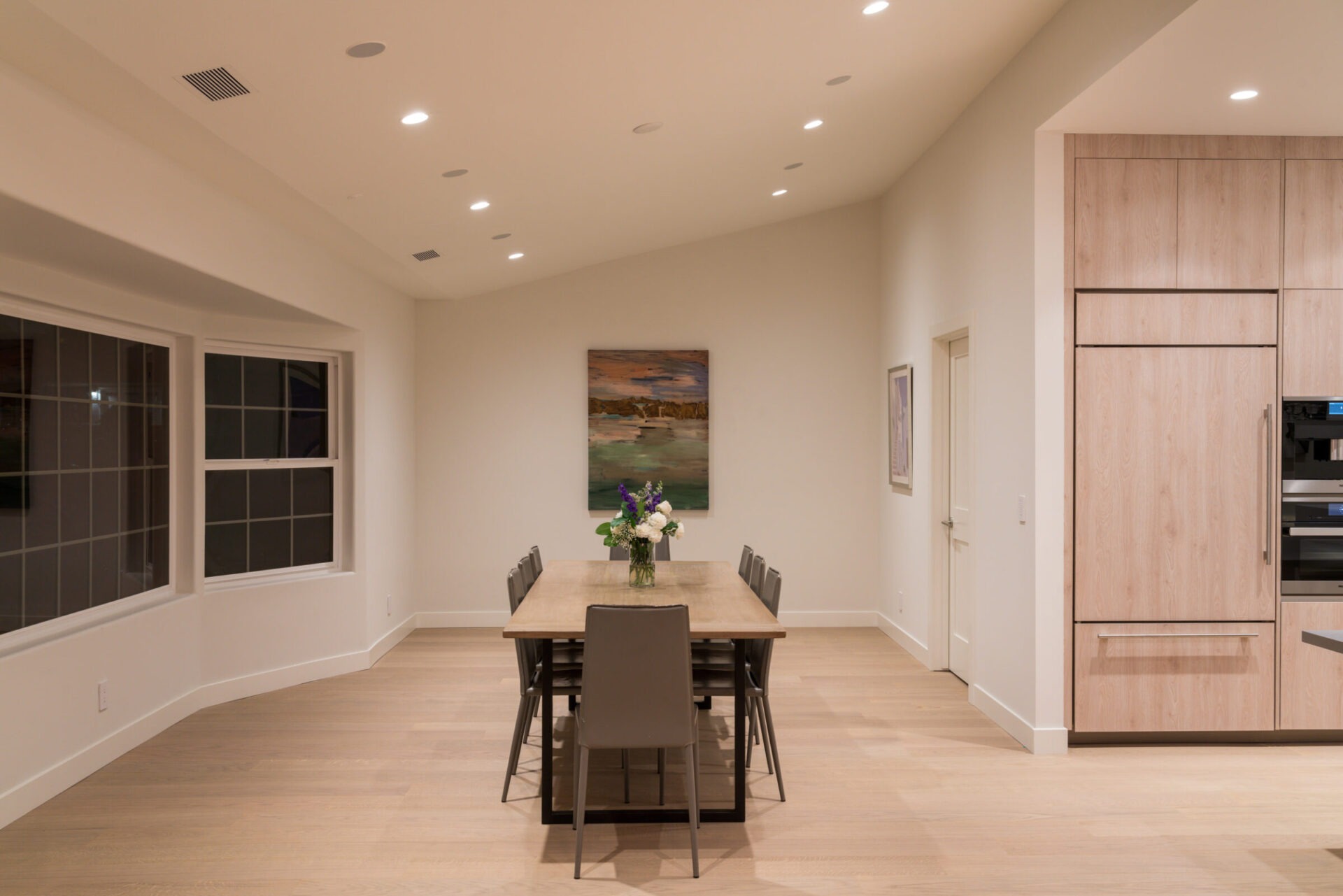 Minimalist dining room with a long wooden table, six chairs, and a vase of flowers. Bright lighting and neutral decor add warmth.