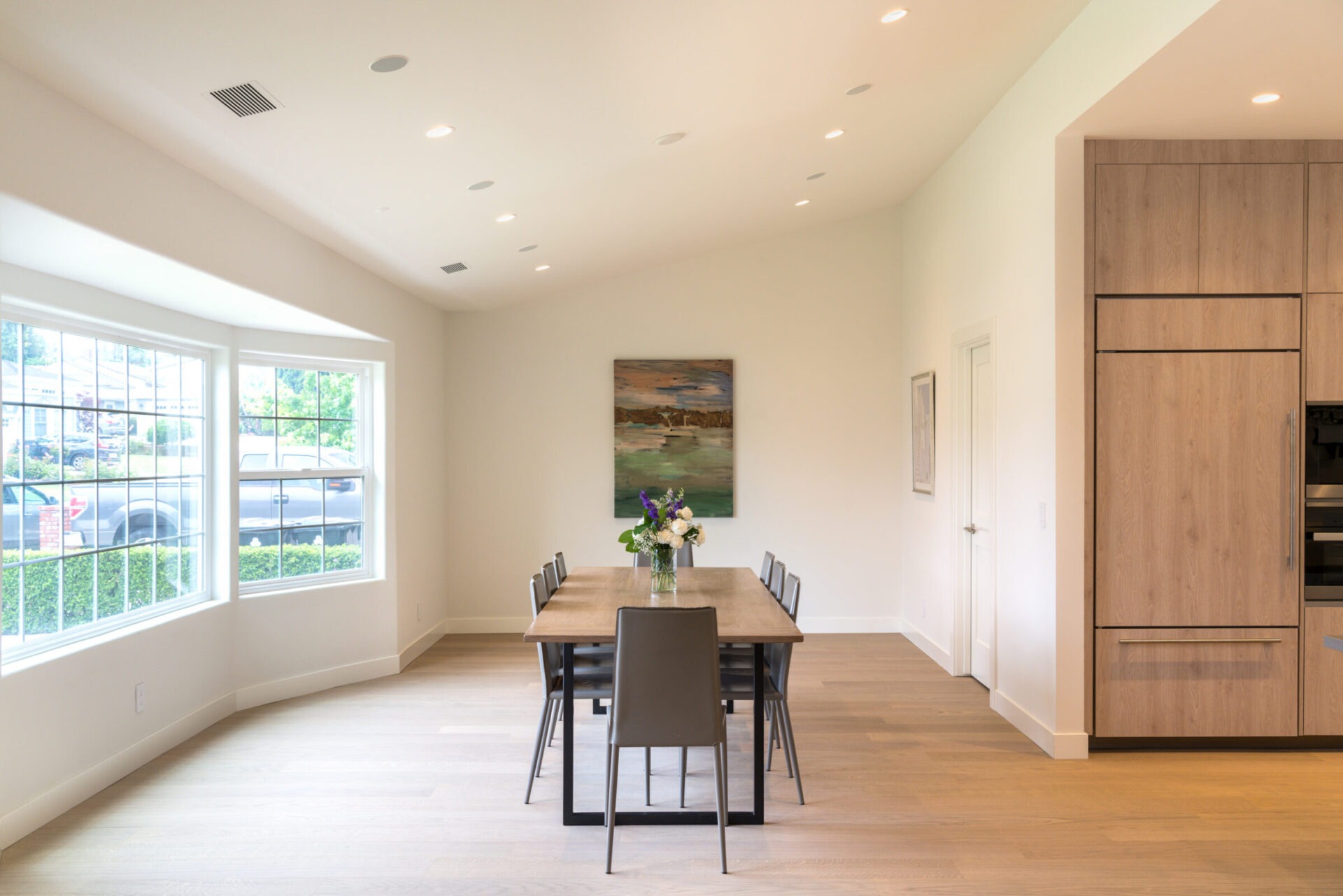 Minimalist dining room with a wooden table, six chairs, and a floral centerpiece. Large windows offer natural light and a scenic view.