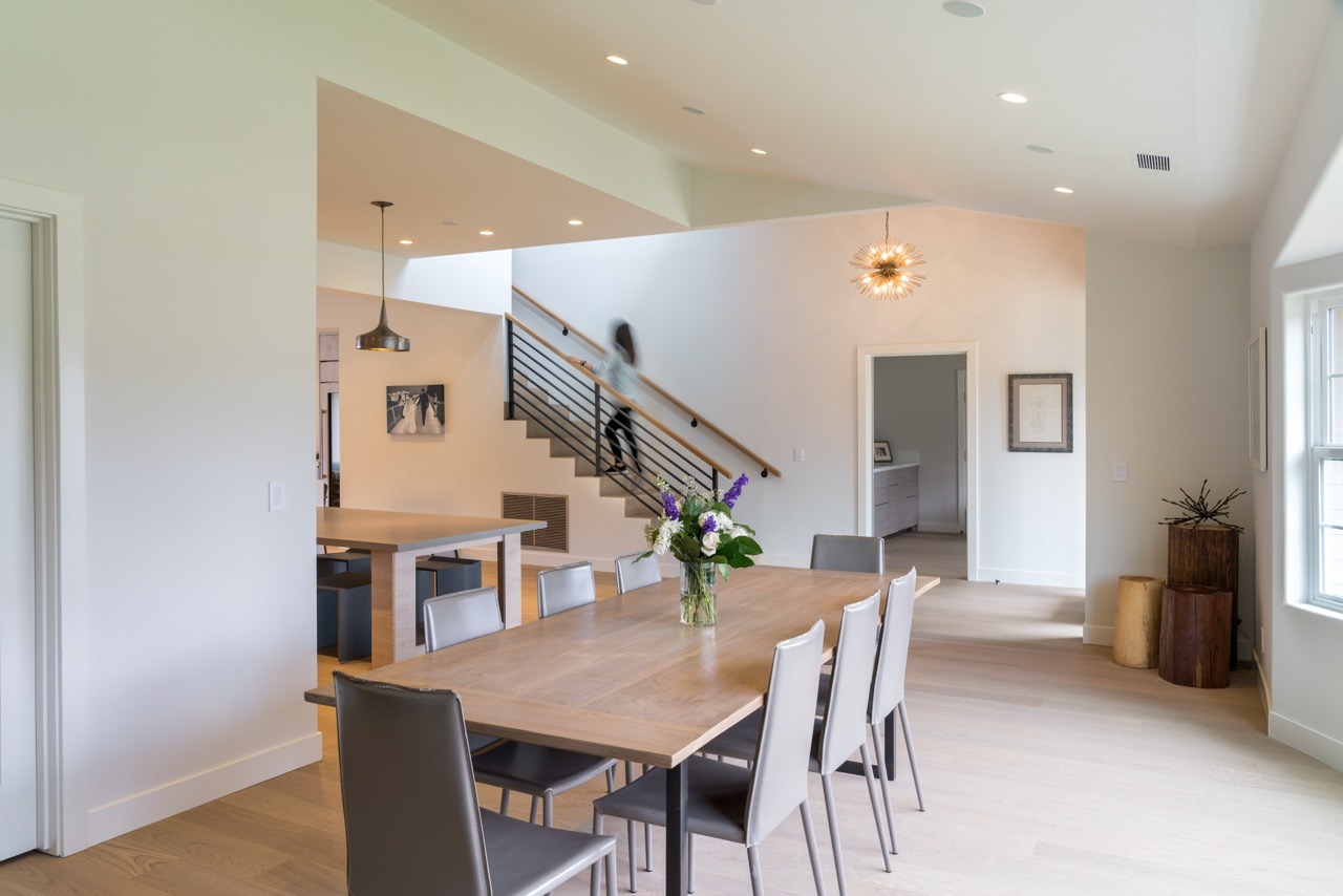 Modern dining area with a wooden table and chairs, staircase, and a person in motion. Bright space with minimalist decor and lighting.