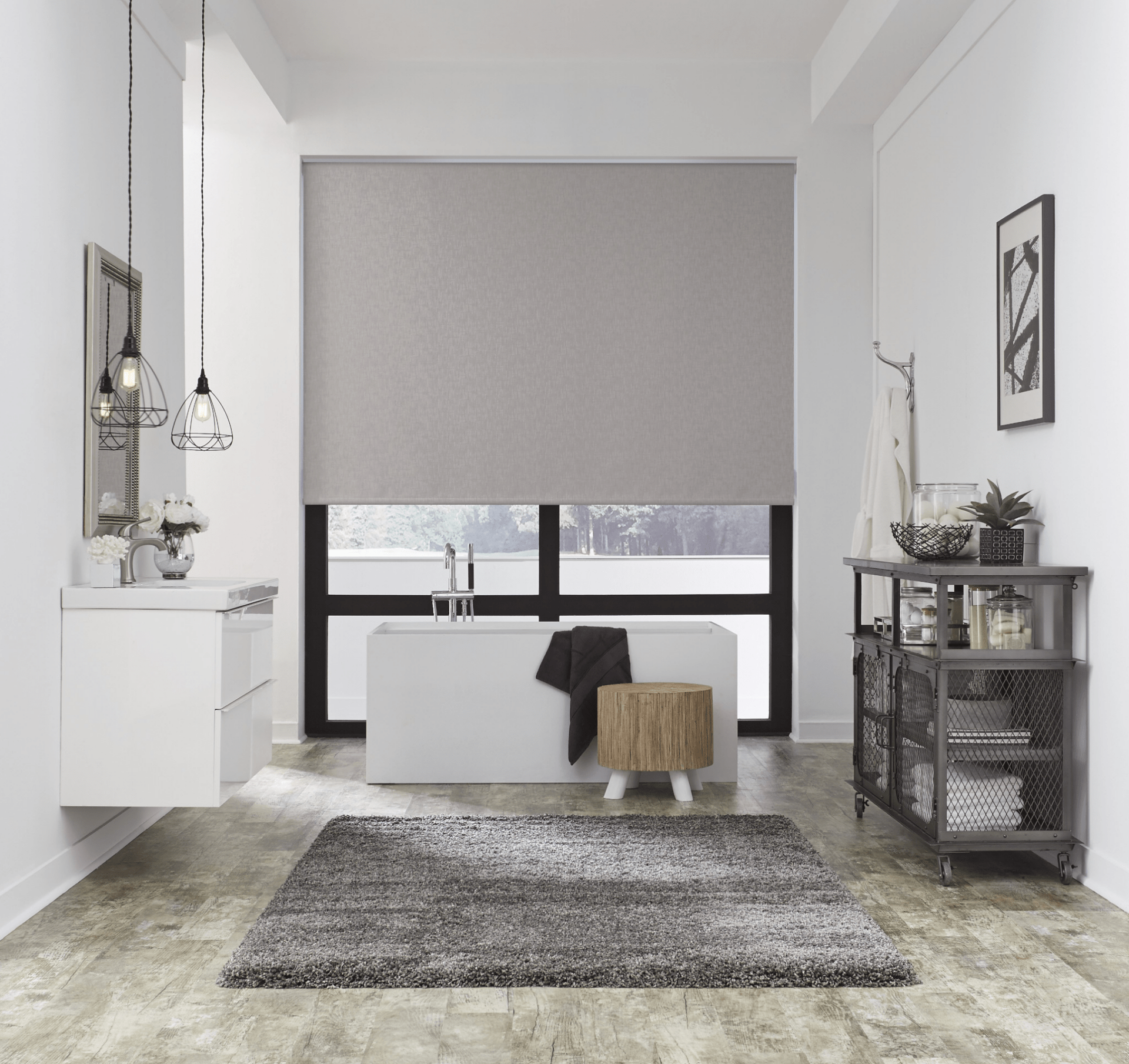 Modern bathroom with a freestanding tub, rug, and minimalist decor. Large window with a roller blind, pendant lighting, and a metal storage unit.