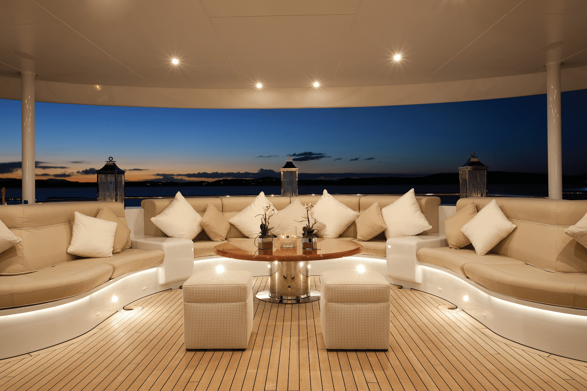 Luxurious yacht deck at sunset, featuring elegant seating with white cushions and soft lighting, overlooking a serene water horizon with painted skies.