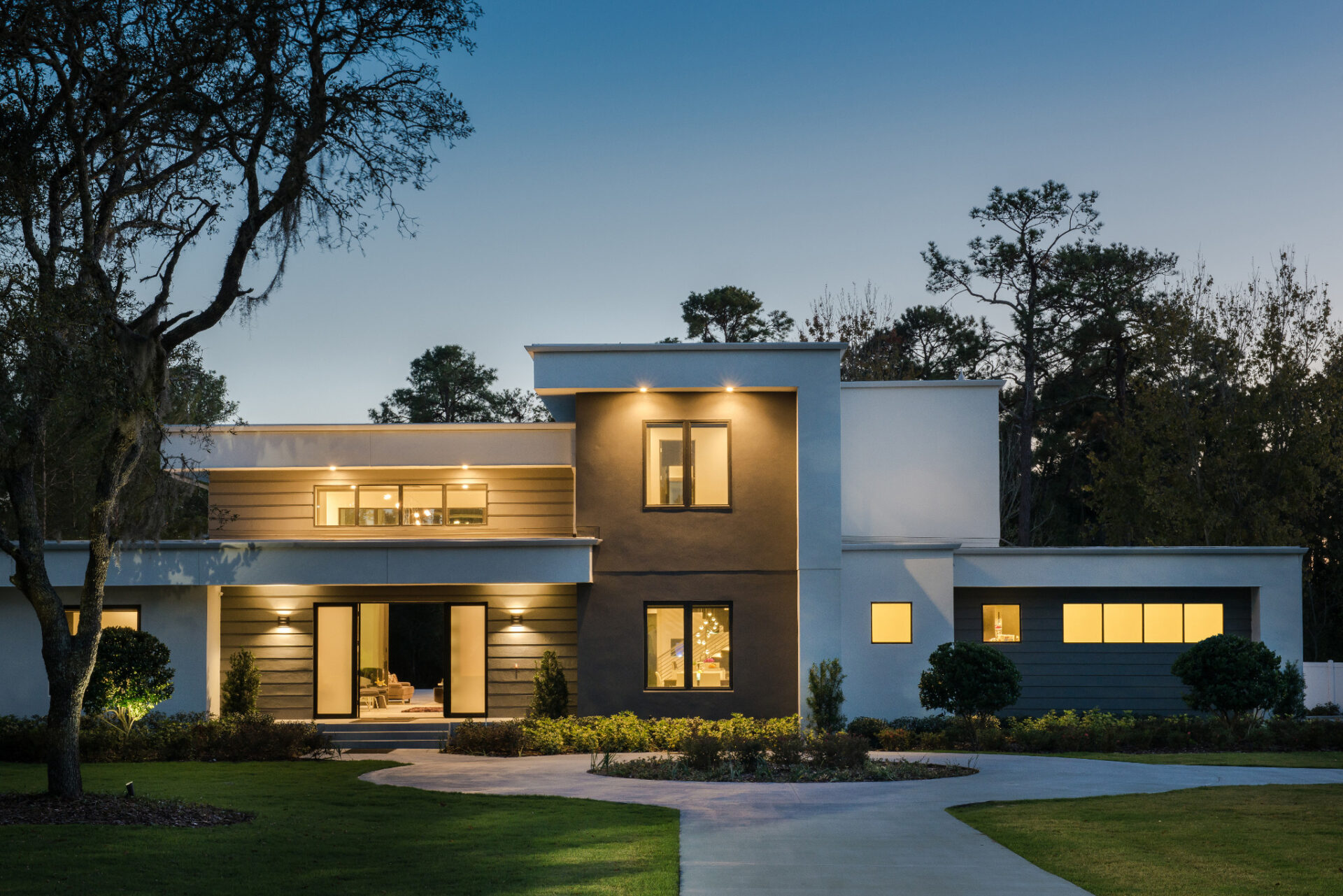 Modern house at dusk with large windows and illuminated interiors. Surrounded by trees and a curved driveway, set against a twilight sky.