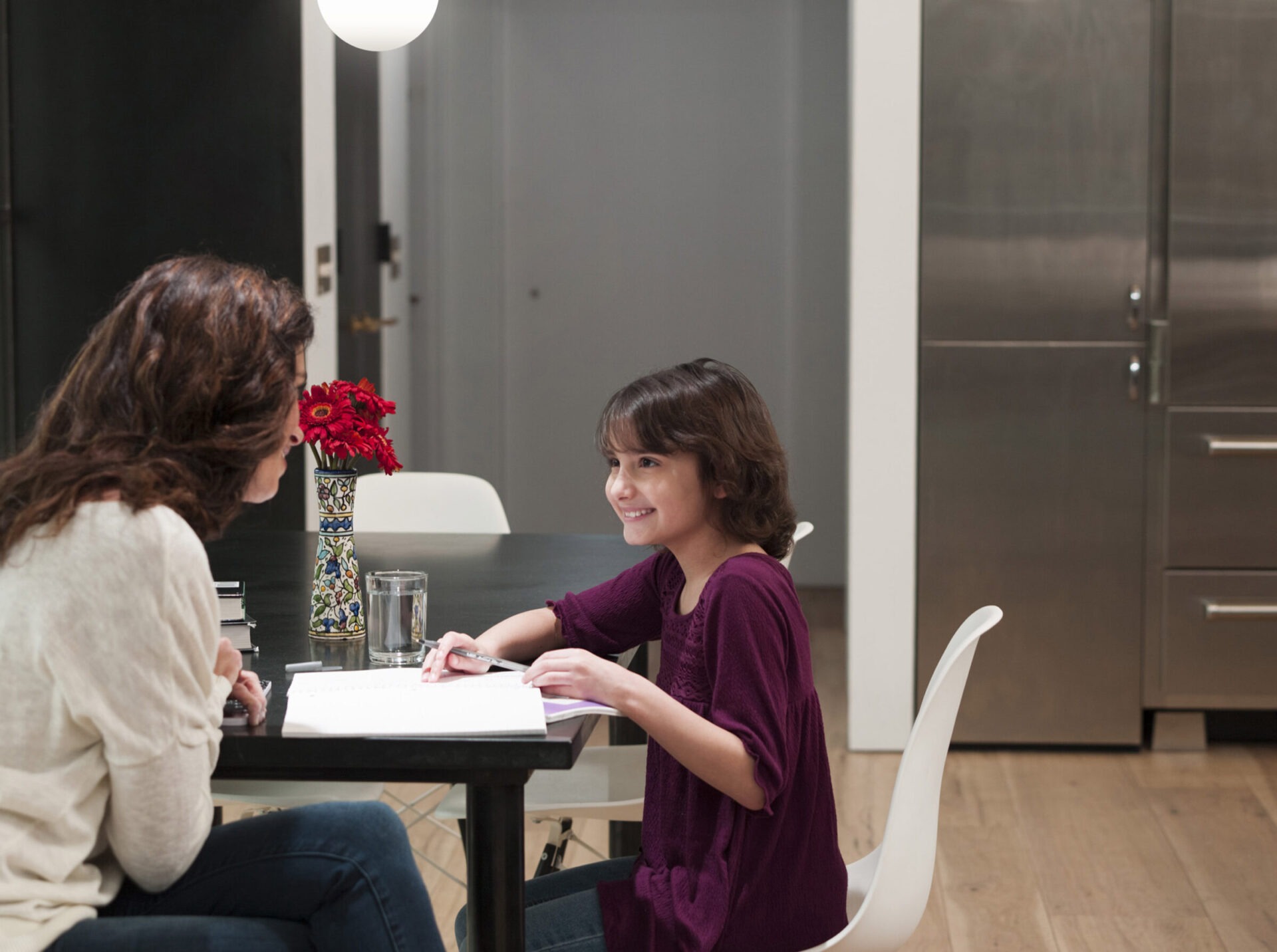 A person and a child are sitting at a dining table, engaging in conversation, with notebooks and a vase of flowers present.