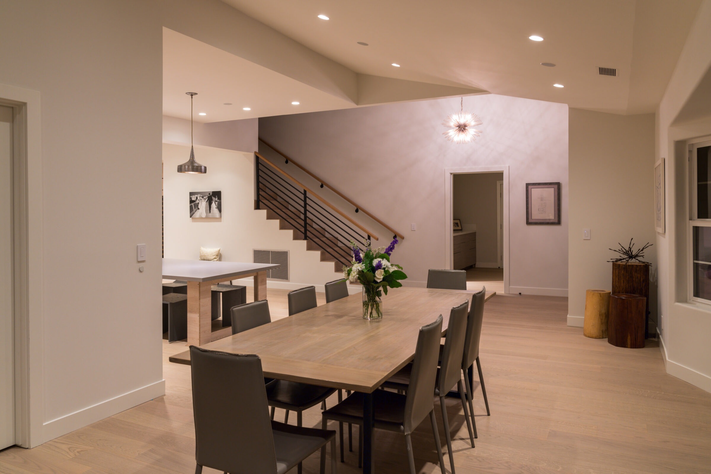 A modern dining area features a long table with chairs, minimalist decor, a staircase, and soft lighting, creating a cozy atmosphere.