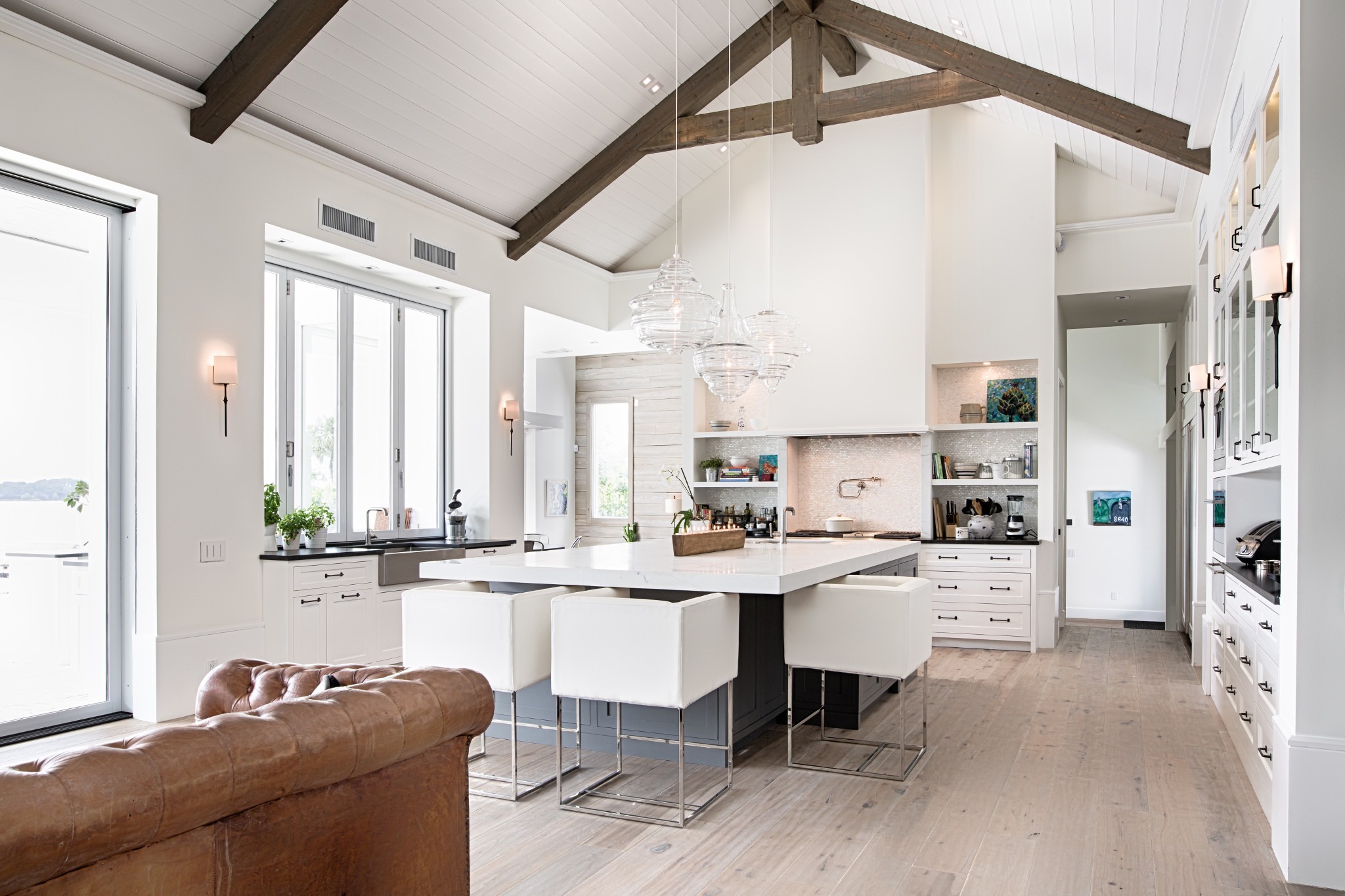 Bright, modern kitchen with high ceilings, exposed beams, large island, white cabinets, and leather couch. Open shelving displays decor. Cozy and stylish atmosphere.