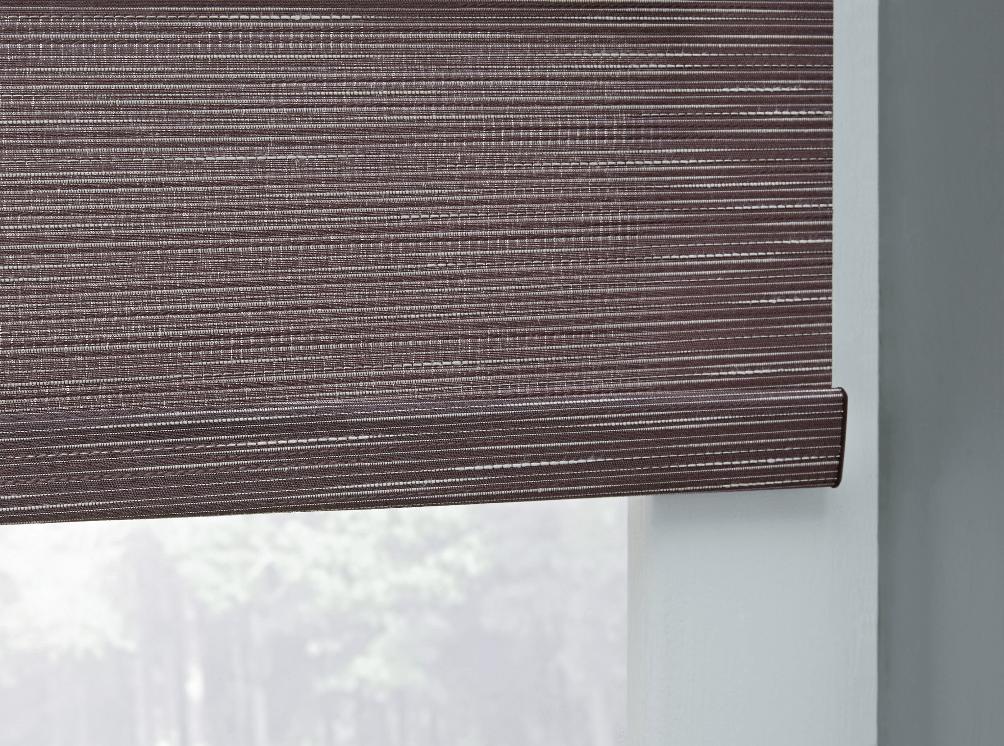 A window with a brown textured roller blind partially drawn, revealing a blurry outdoor view. Natural light filters through, creating a serene atmosphere.