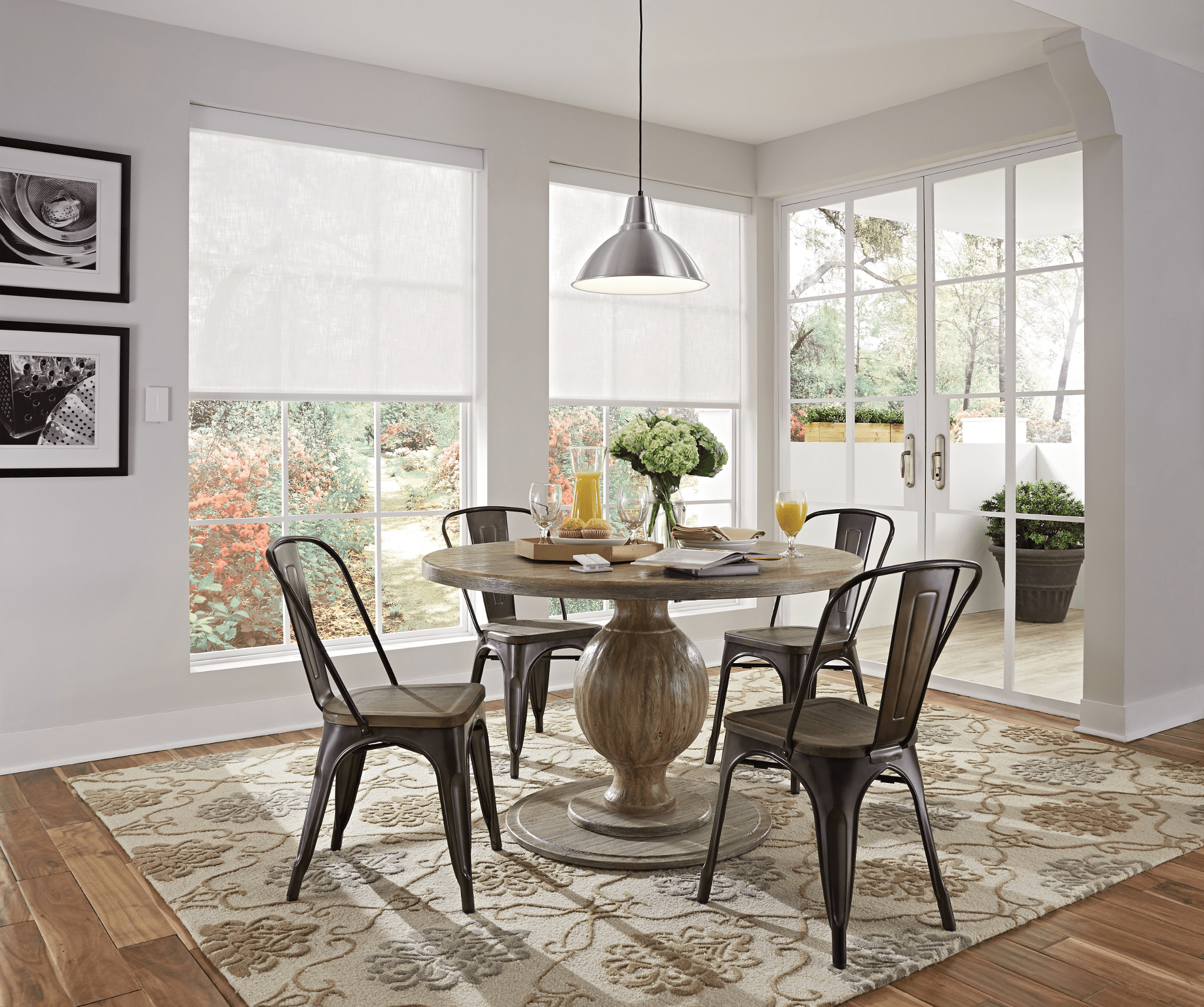 Bright dining room with round table, metal chairs, pendant light, and decorative rug. Large windows showcase a garden view with vibrant foliage.