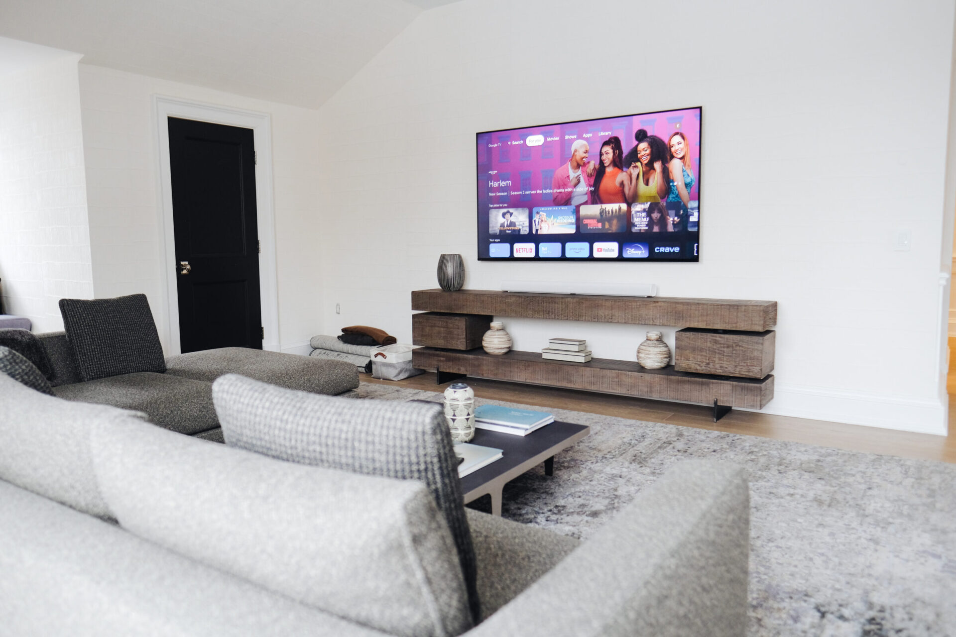 A modern living room features a large wall-mounted TV, gray sofas, and a wooden entertainment unit with decorative vases and books.