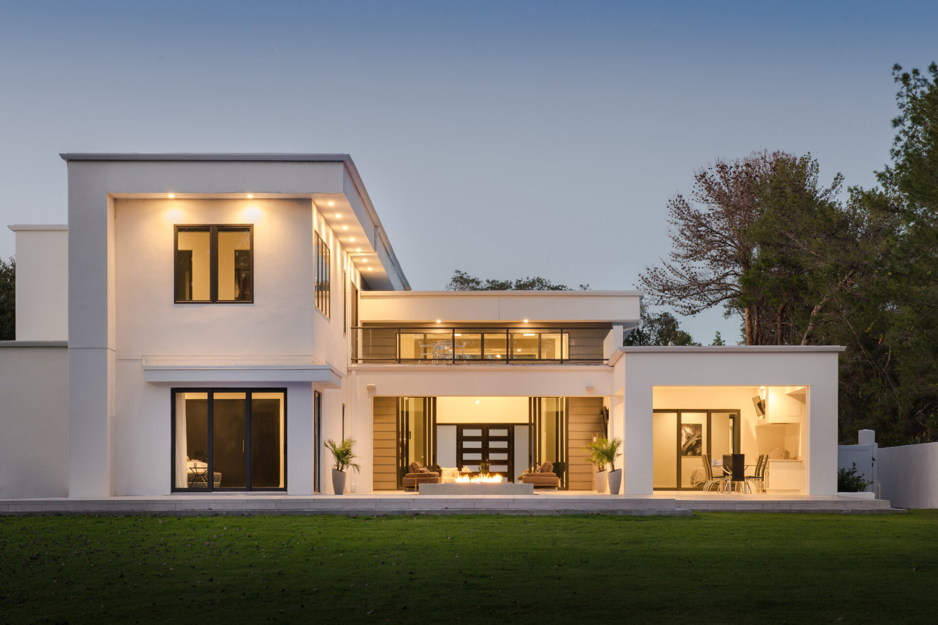 Modern two-story white house with large windows and outdoor patio area, surrounded by a well-kept lawn and trees in the background.