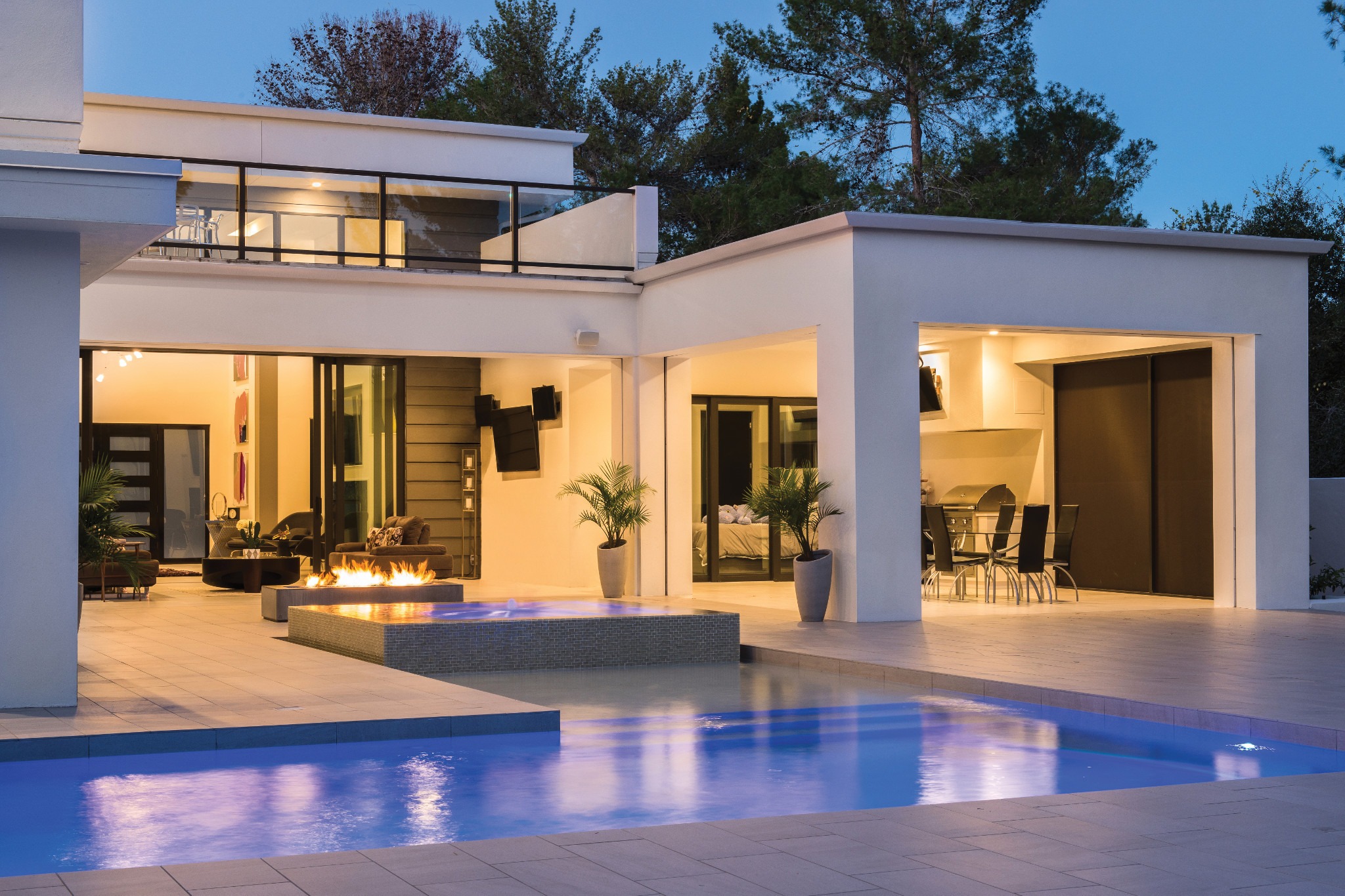 Modern house with large windows, outdoor pool, and fire pit at dusk. Surrounded by trees, featuring minimalistic decor and open spaces for relaxation.