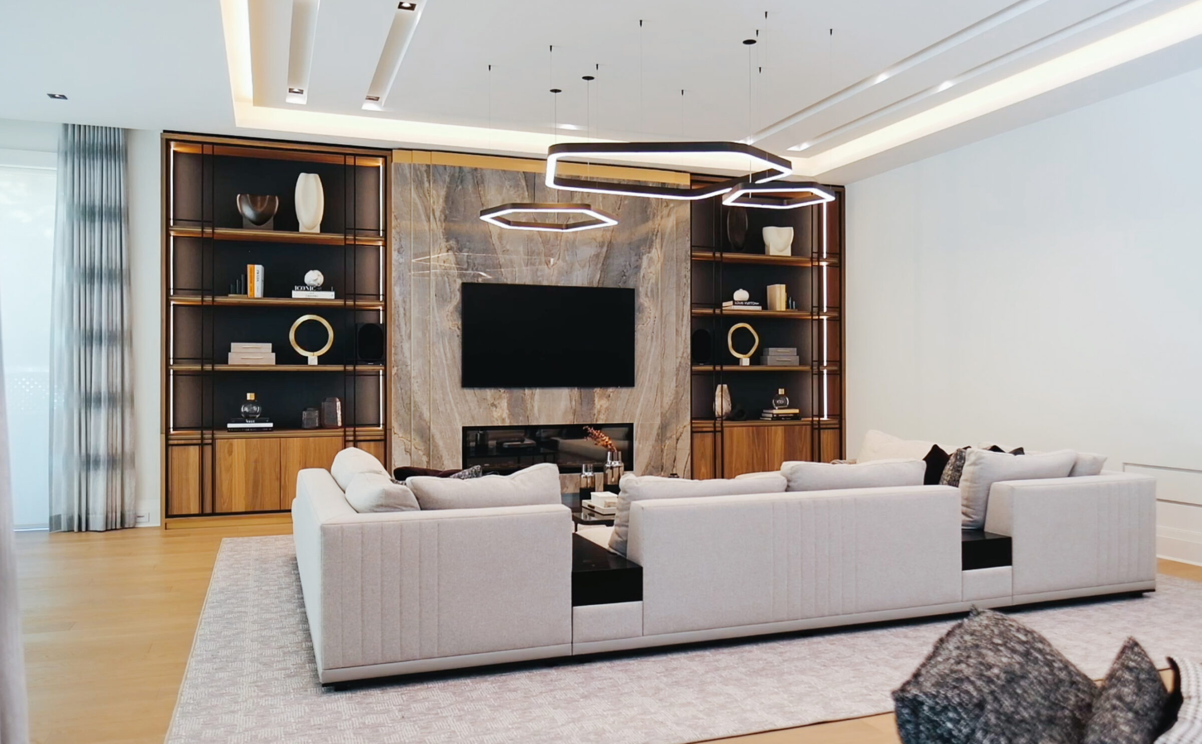 Modern living room with a large sectional sofa, elegant shelving, abstract lighting fixtures, and a mounted TV on a marble accent wall. Cozy and stylish ambiance.