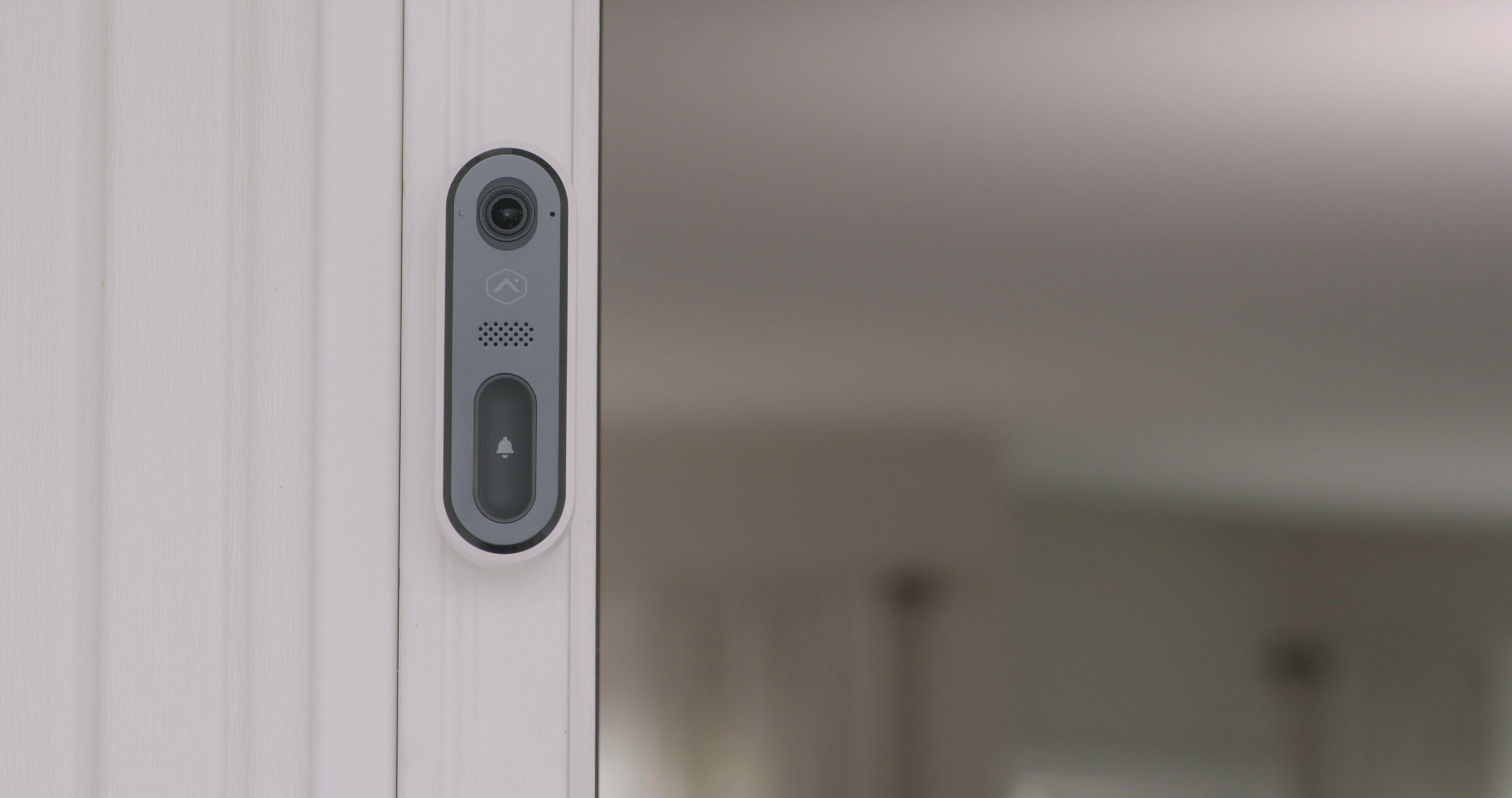 A modern doorbell camera is mounted on a white door frame, inside a brightly lit, blurred interior setting. No landmarks or people are visible.