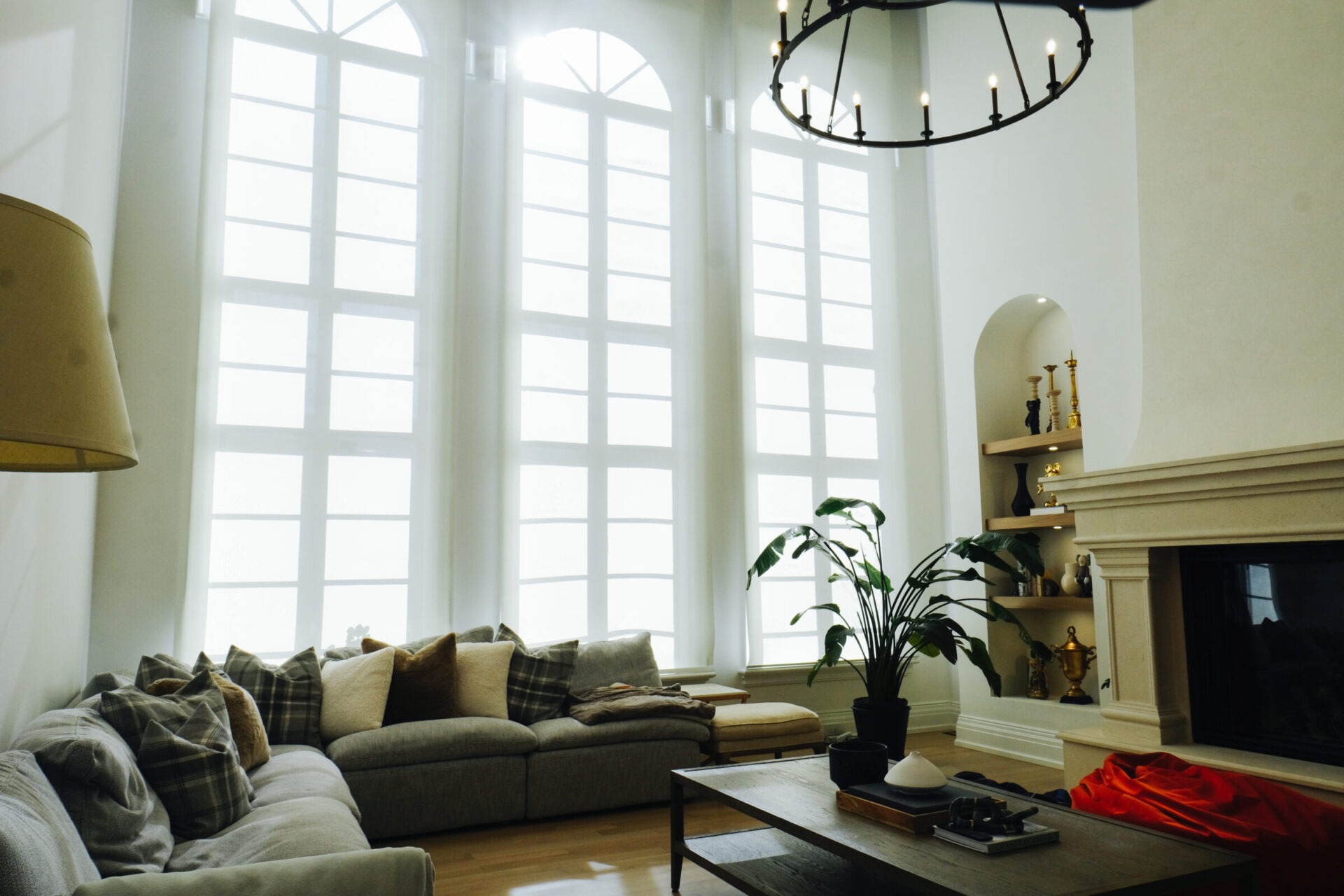 Cozy living room with tall arched windows, elegant chandelier, comfortable sofa, and a fireplace. Decor includes a plant and colorful accents on shelves.