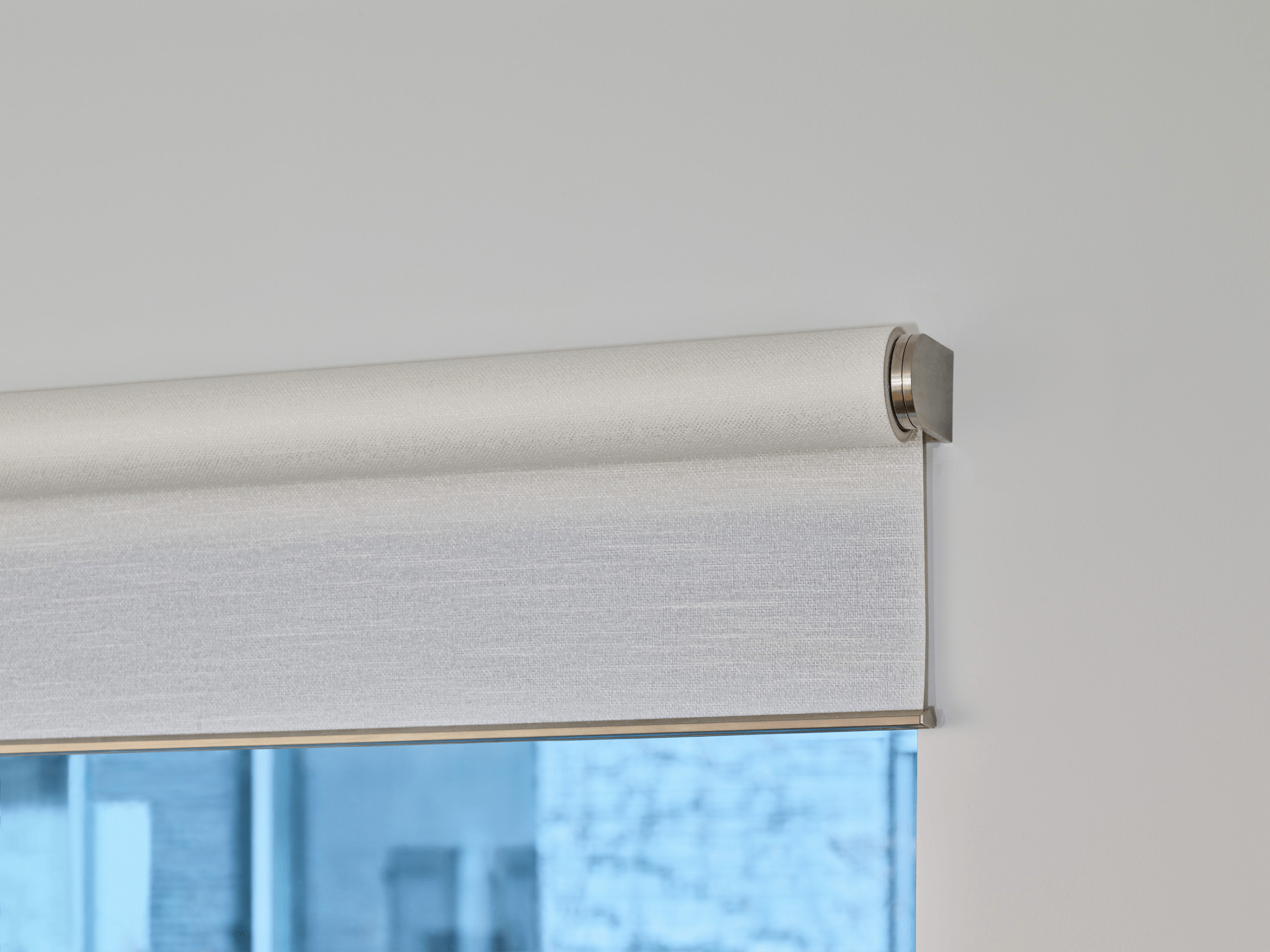 A close-up of a modern roller blind partially covering a window, with a blurred urban building visible in the background.