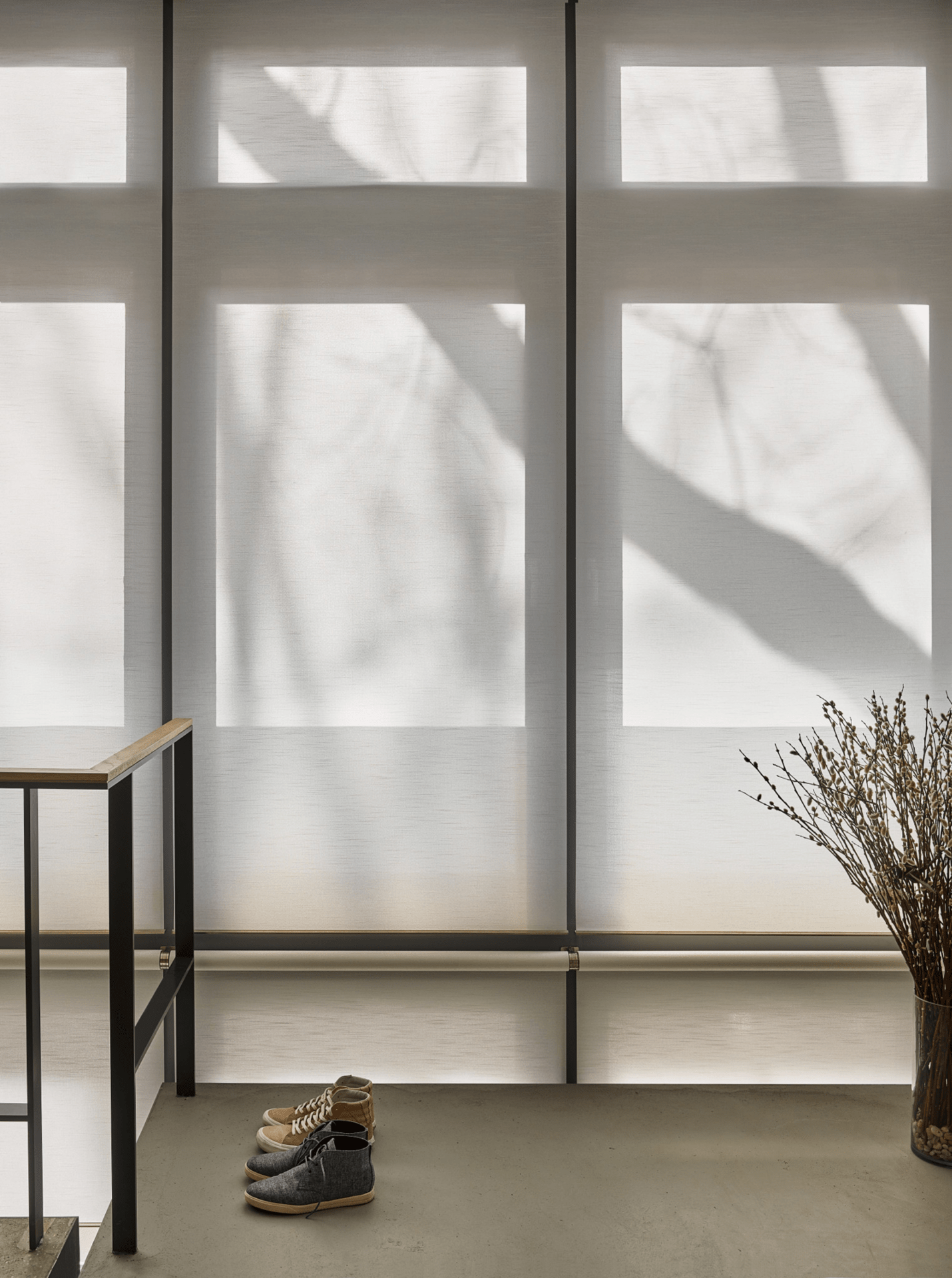 A serene interior features large windows with tree shadows, a pair of boots, and a potted plant beside a modern railing.