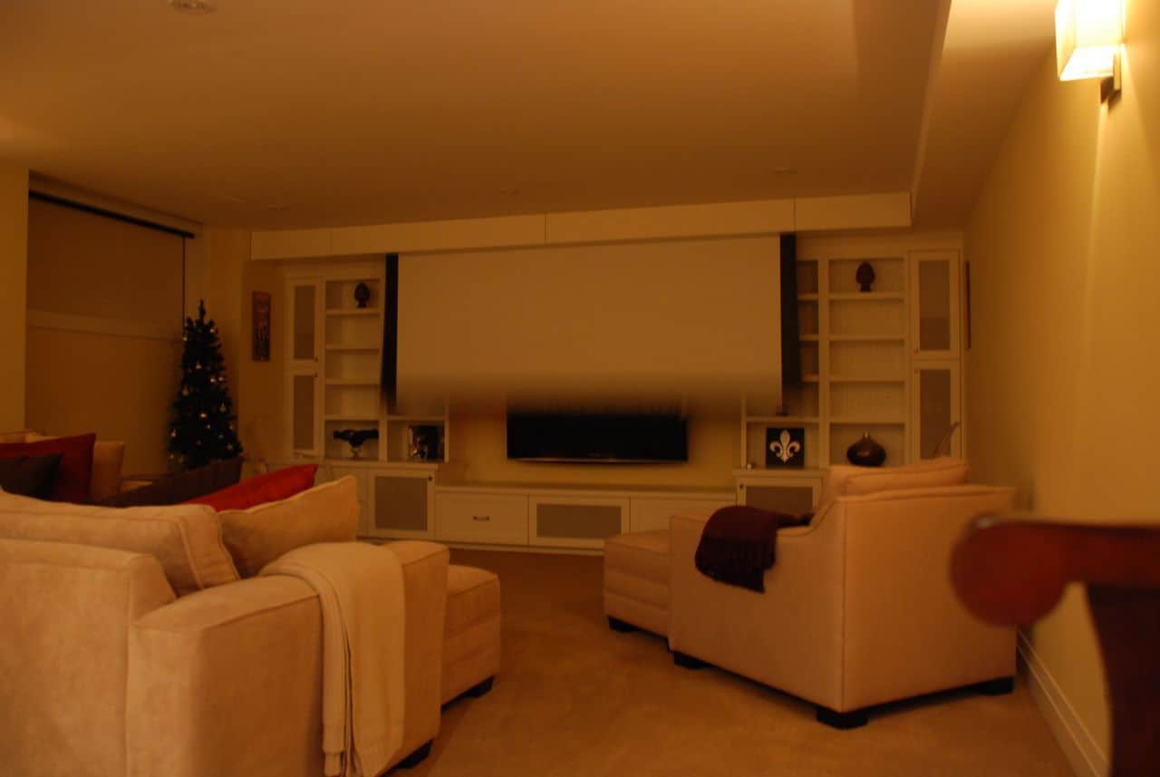 Cozy living room with beige sofas, a large TV, bookshelves, dim lighting, and a decorated Christmas tree in the corner.