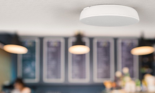 A white smoke detector is mounted on a ceiling in a café, with blurred menu boards and hanging lights in the background.