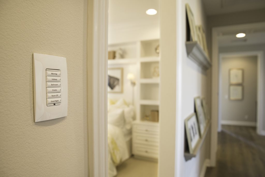 A modern hallway with smart light control panel, leading to a neatly decorated bedroom with shelves and framed pictures on the wall.