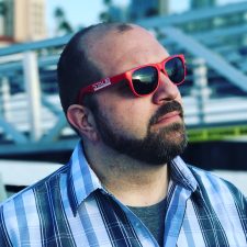 A person wearing red sunglasses and a plaid shirt gazes upwards. The background includes metal structures and palm trees under a clear sky.