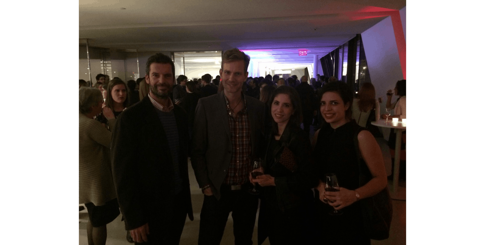 A group of people at a lively indoor gathering, casually dressed, holding drinks. The background has dim lighting and an illuminated ceiling.