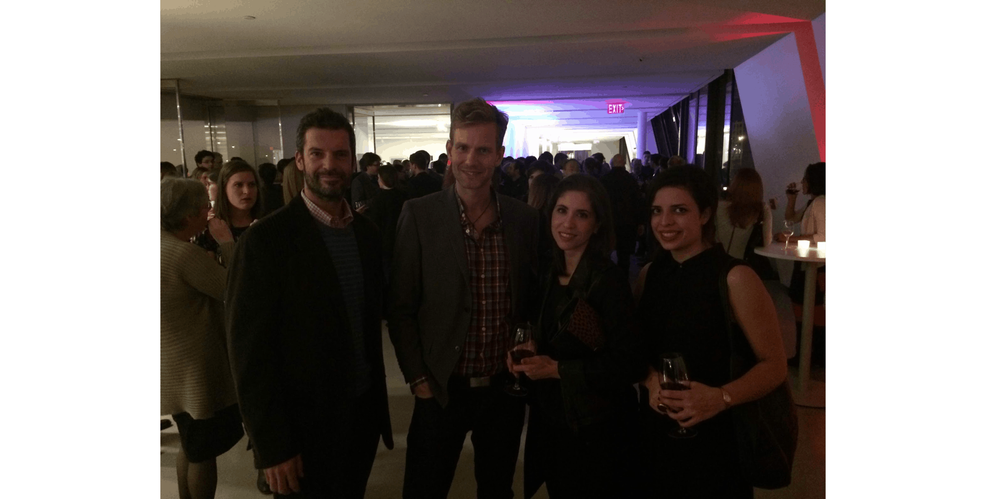 A group of people at a lively indoor gathering, casually dressed, holding drinks. The background has dim lighting and an illuminated ceiling.