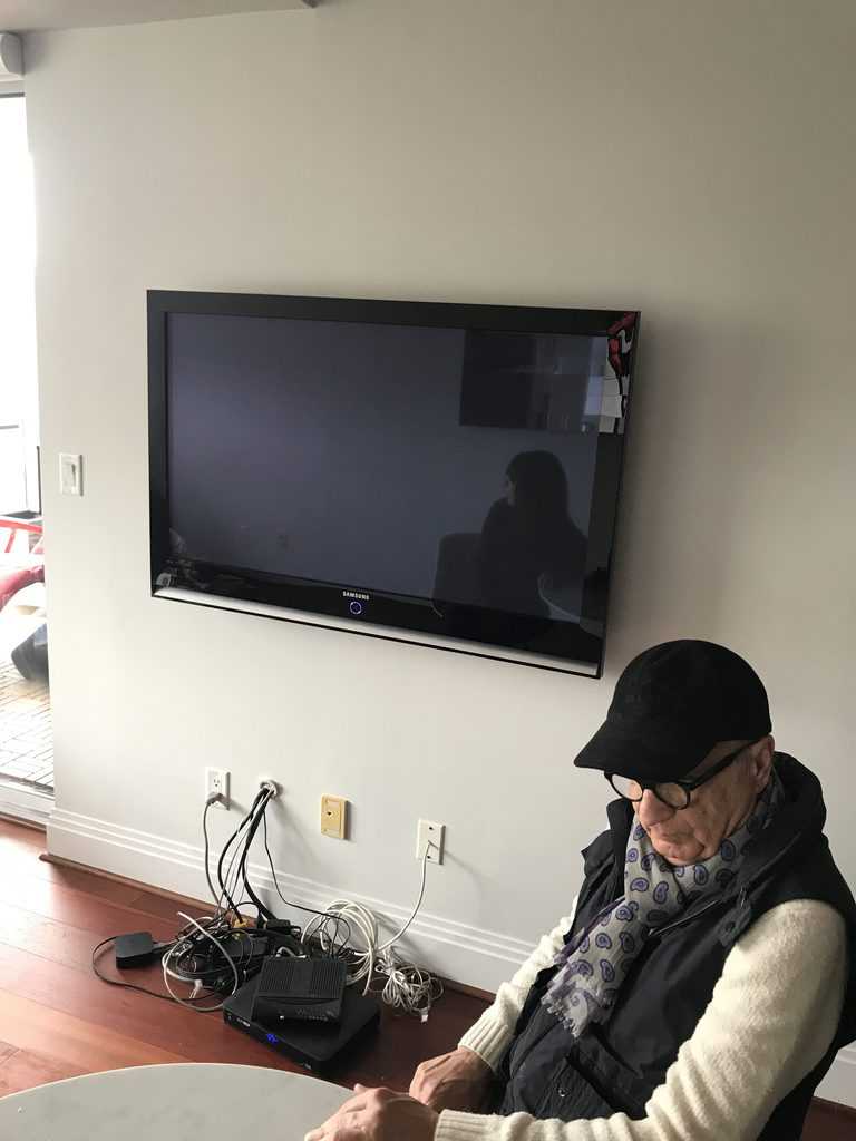 A person sits beside a wall-mounted TV with visible cables and equipment below. A reflection is seen on the screen, and sunlight enters.