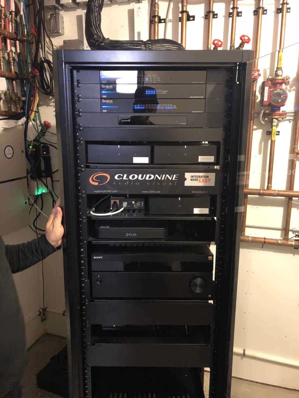 A person stands next to a server rack filled with various audio-visual equipment, including a Cloud9 Audio Visual device, in a utility room.