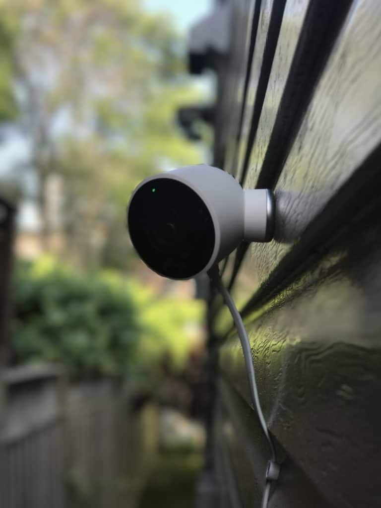 A wall-mounted security camera focuses on an outdoor space, with trees and a blurred background visible, suggesting a residential setting.