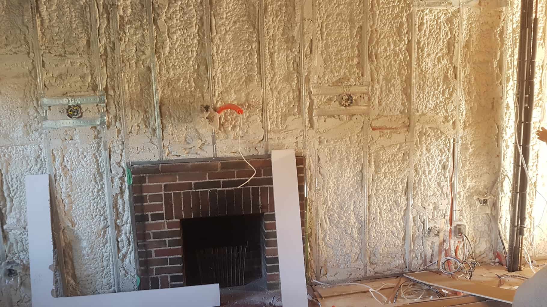 Room with exposed insulation and brick fireplace, wires hanging down. Foam insulation is applied on the walls, with construction materials scattered around.