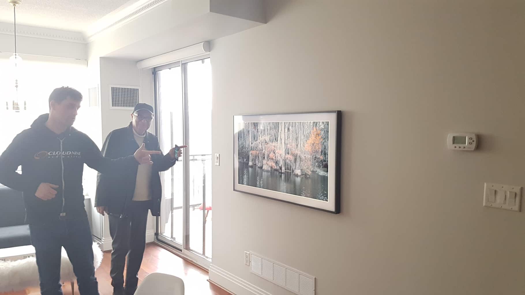 Two people are indoors, examining a framed photo on the wall. Natural light enters through a sliding door to their right.