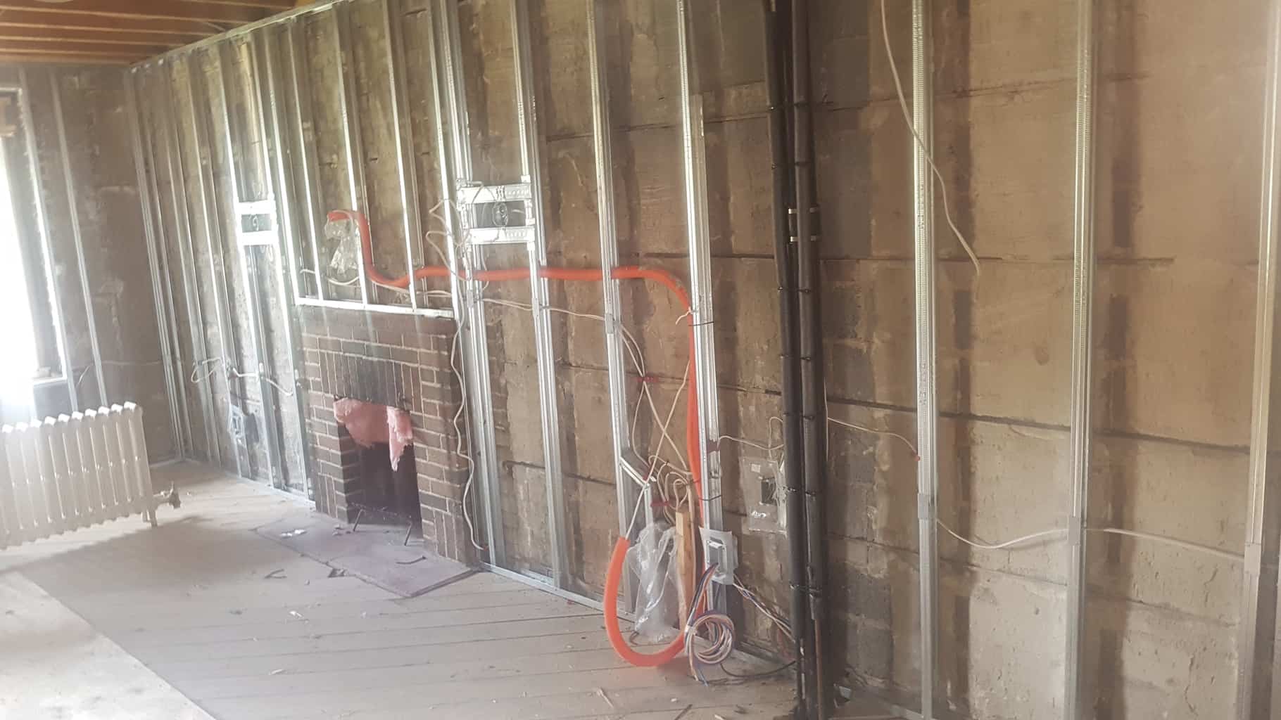 Partially renovated room with exposed stud walls and wiring. Features include a brick fireplace, wooden floor, and a white radiator. No landmarks visible.