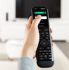 A person holds a Logitech remote, pointed towards a blurred television, in a modern, bright living room setting with minimal decor.