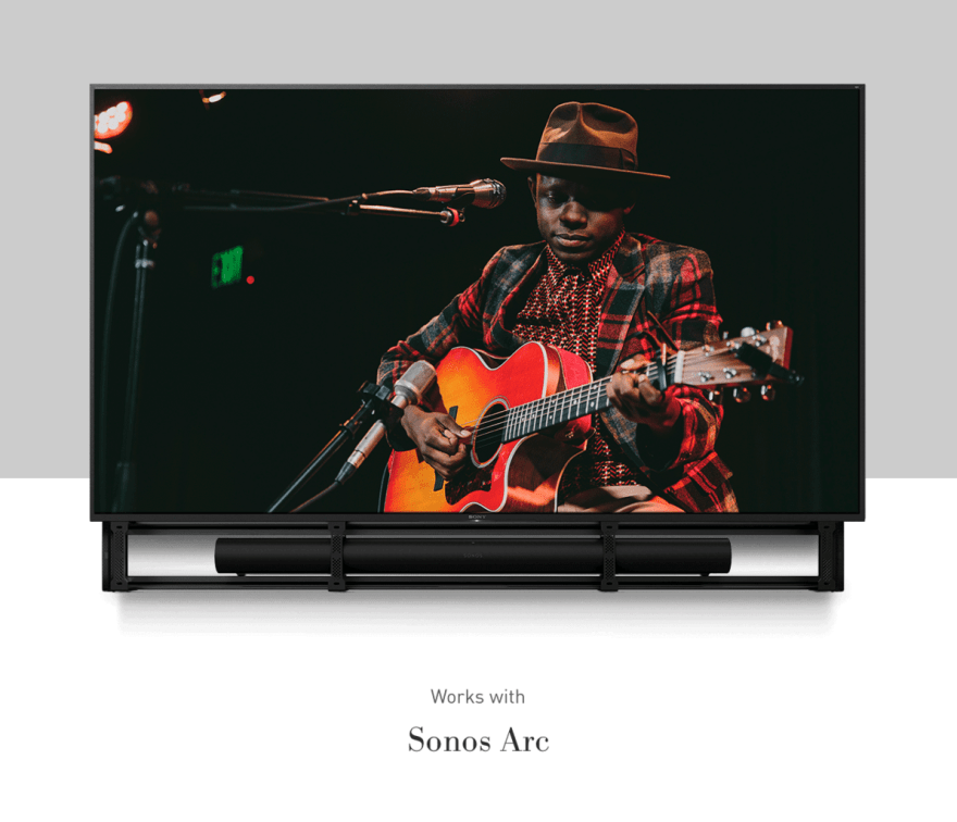 A person in a plaid jacket plays guitar on stage, displayed on a screen above a Sonos Arc soundbar system.