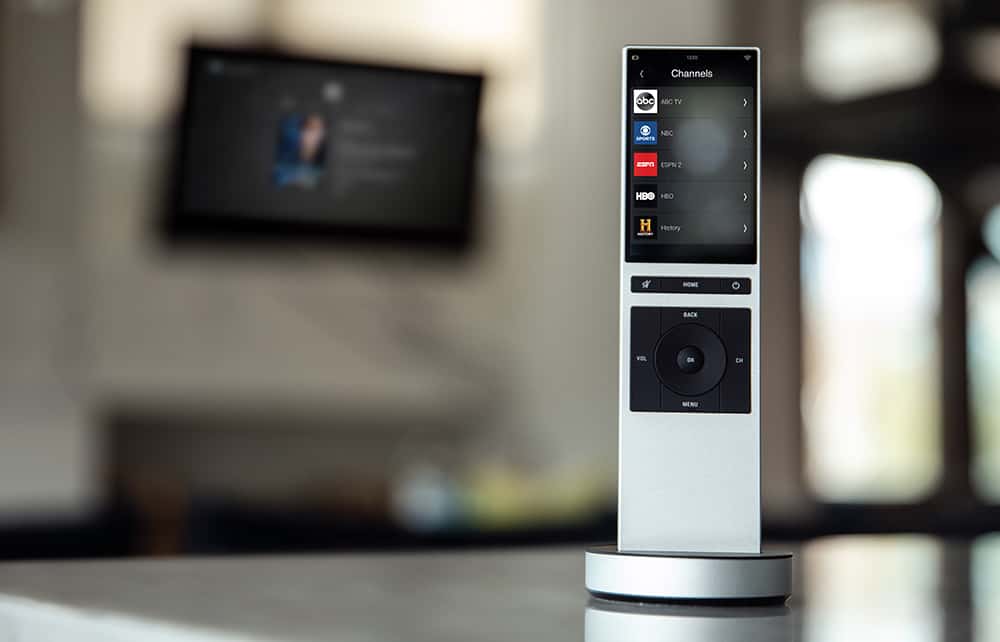 A modern smart remote control stands upright on a table in a well-lit room, with a blurred television screen in the background.