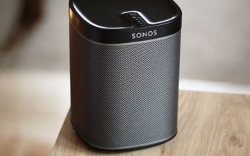 A Sonos wireless speaker is placed on a wooden surface, with no visible background details or people in the image.