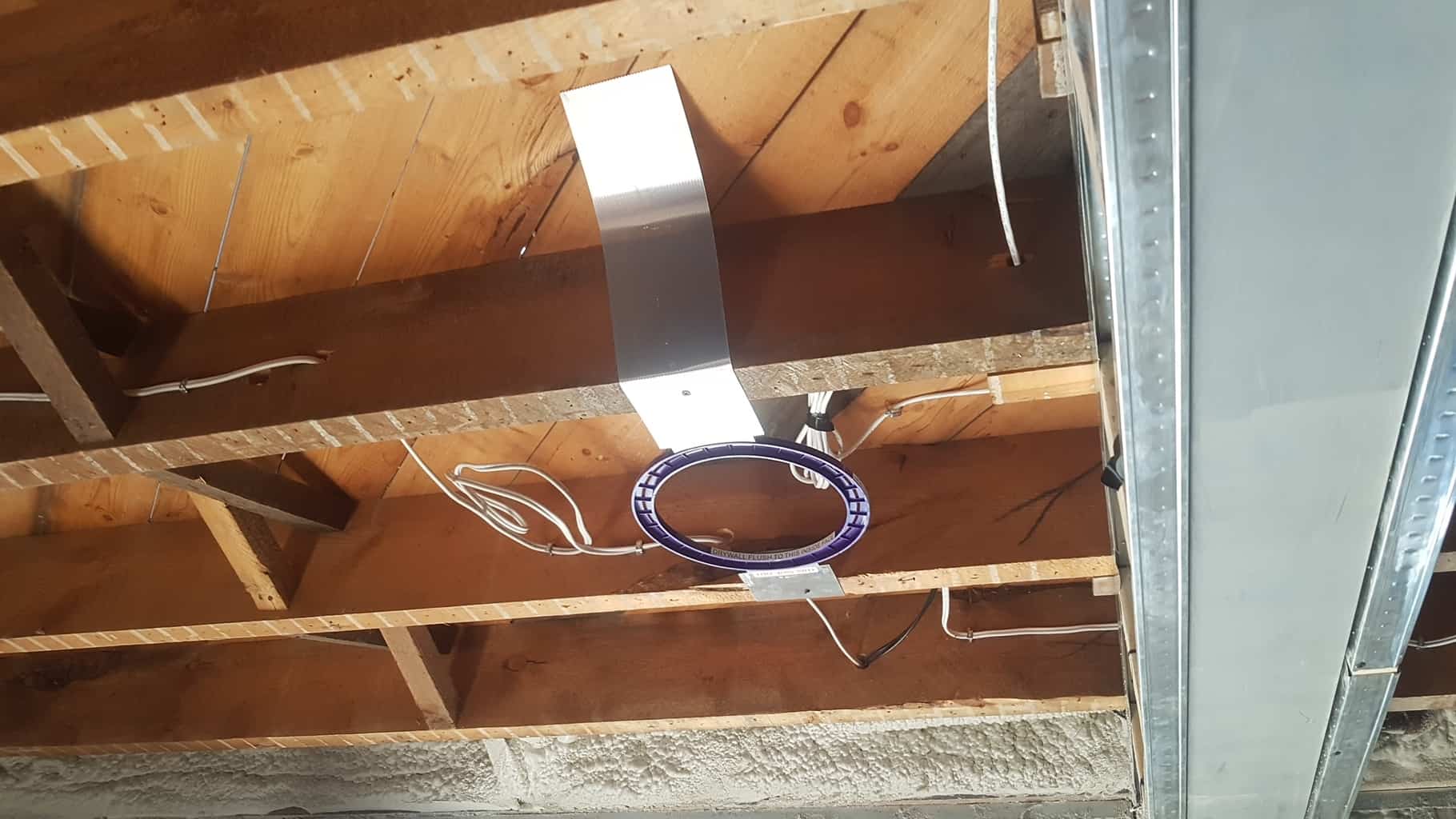 An unfinished ceiling with exposed wooden beams and electrical wires. A circular device is attached underneath the beams. Industrial and minimalistic setting.