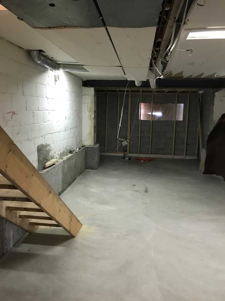 An unfinished basement with exposed ceiling wires, concrete walls, and wooden stairs. A small window provides minimal natural light in the space.