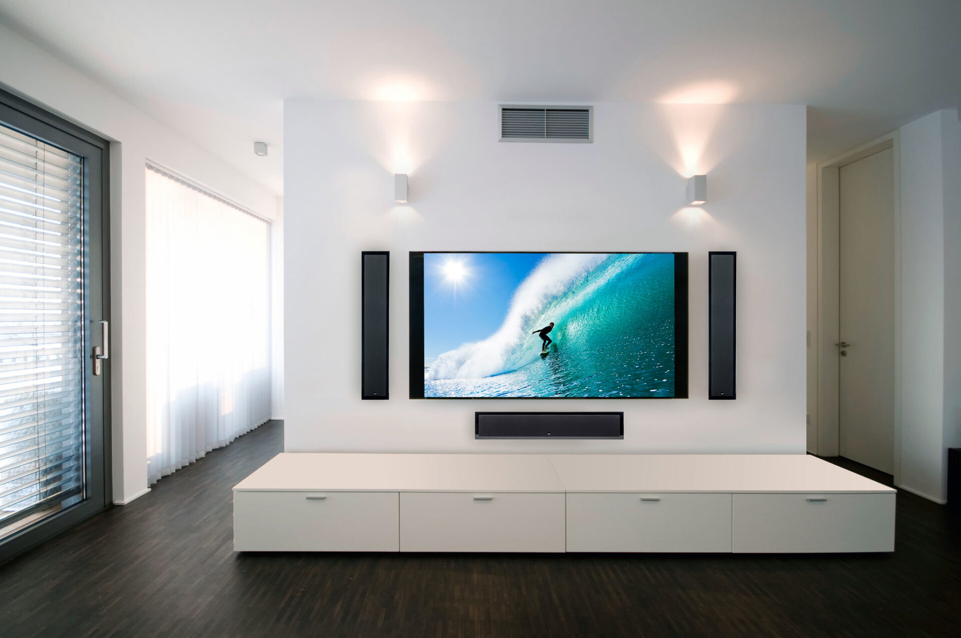 Modern living room with sleek white entertainment unit, large wall-mounted TV showing a surfing scene, and natural light streaming through blinds. Minimalist design.