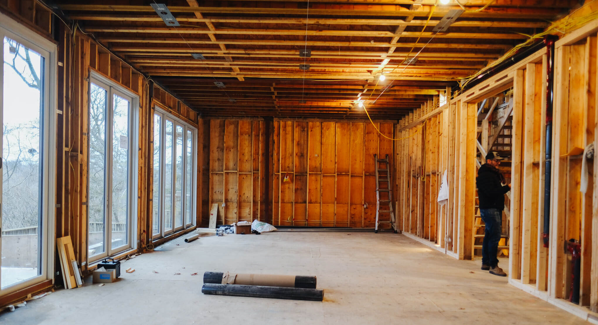 A person stands in a partially constructed room framed with wood. Large windows provide natural light. Construction materials are scattered on the floor.