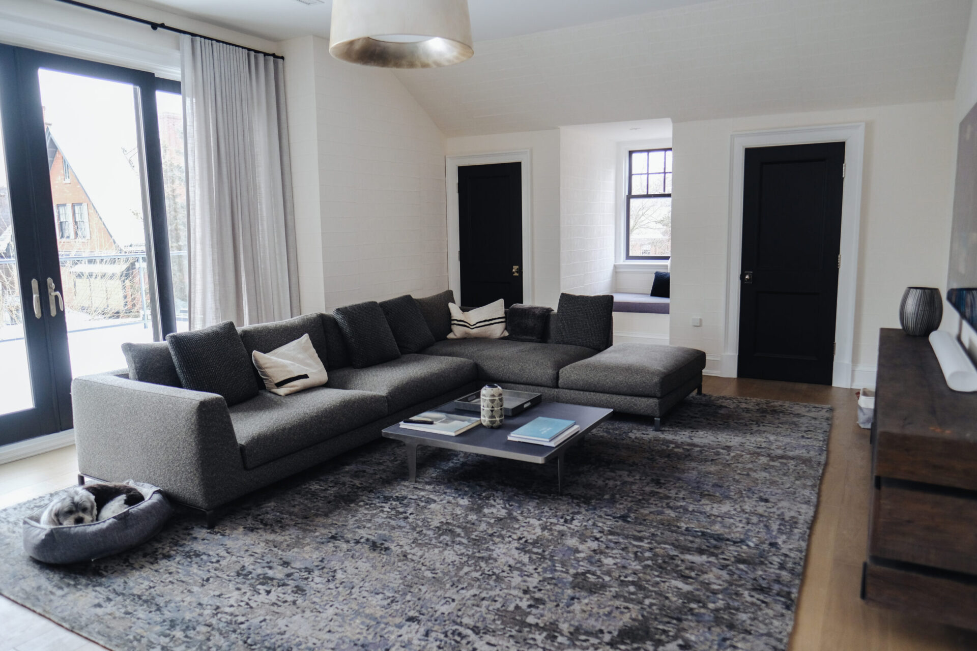 A modern living room features a large gray sectional sofa, coffee table, two doors, and a dog bed on a textured rug.