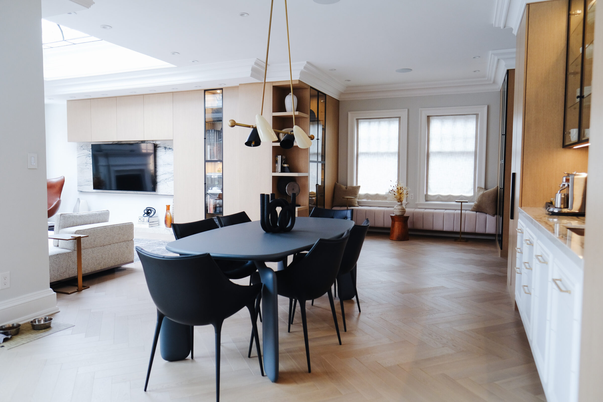 Modern, open-plan living room and dining area featuring minimalist decor, black chairs around a dark table, and a large television opposite a seating area.